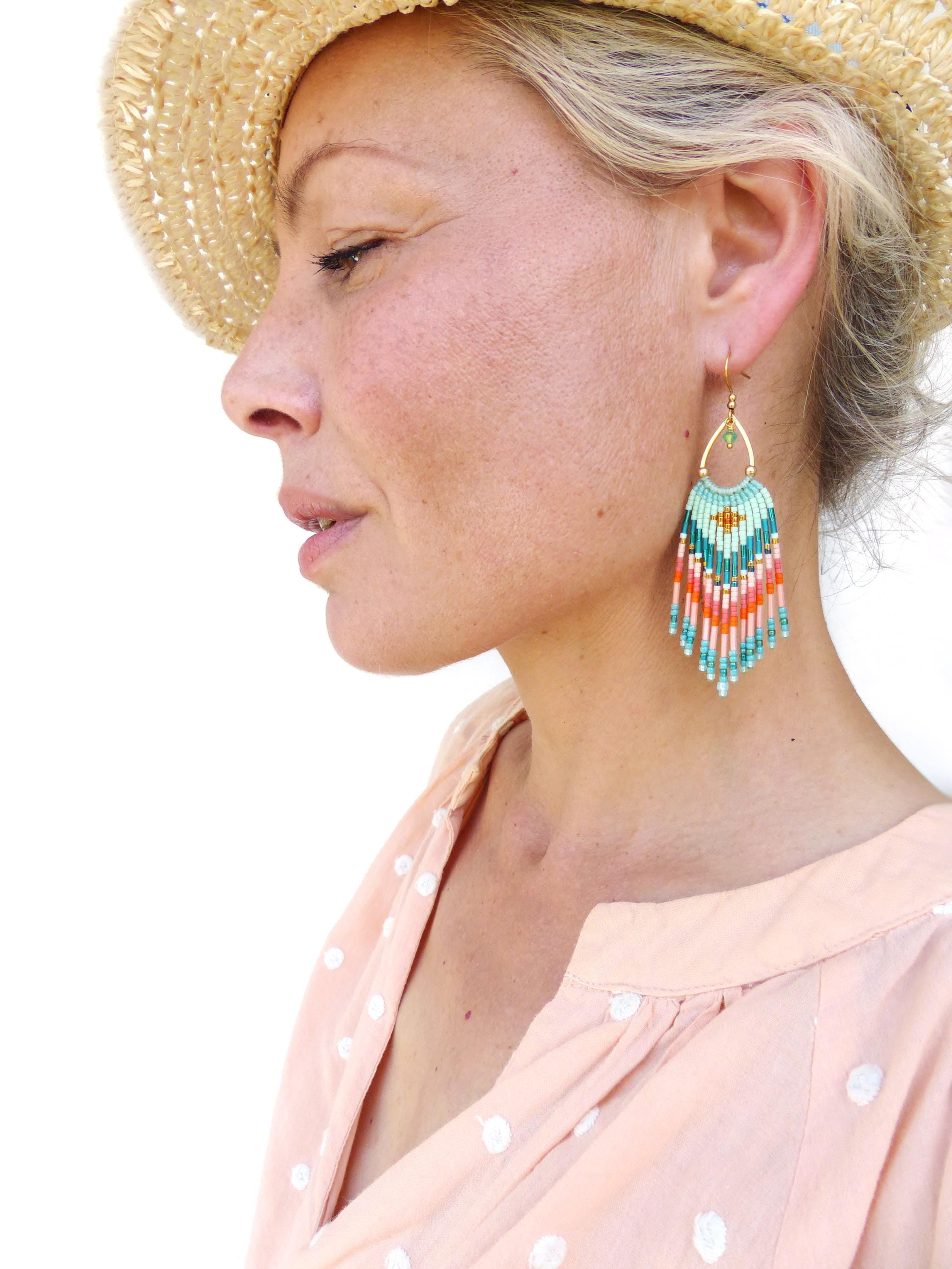 Peut inclure: Une boucle d'oreille pendante composée d'une monture en métal doré en forme de goutte, ornée d'un tissage de perles dans des tons turquoise, corail et blanc. La partie inférieure présente plusieurs rangées verticales de perles assorties.