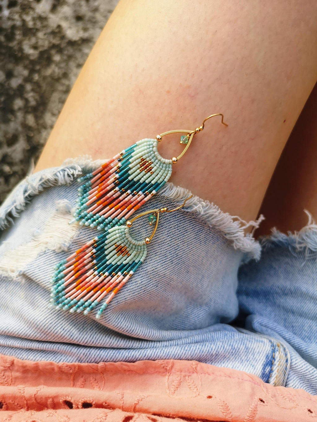 Peut inclure: Une paire de boucles d'oreilles pendantes dotées d'une monture en métal doré en forme de goutte avec un petit cristal vert. Les boucles sont ornées de rangées de minuscules perles en verre bleu clair, corail et bleu sarcelle, disposées en motif géométrique avec une longue frange de perles multicolores.