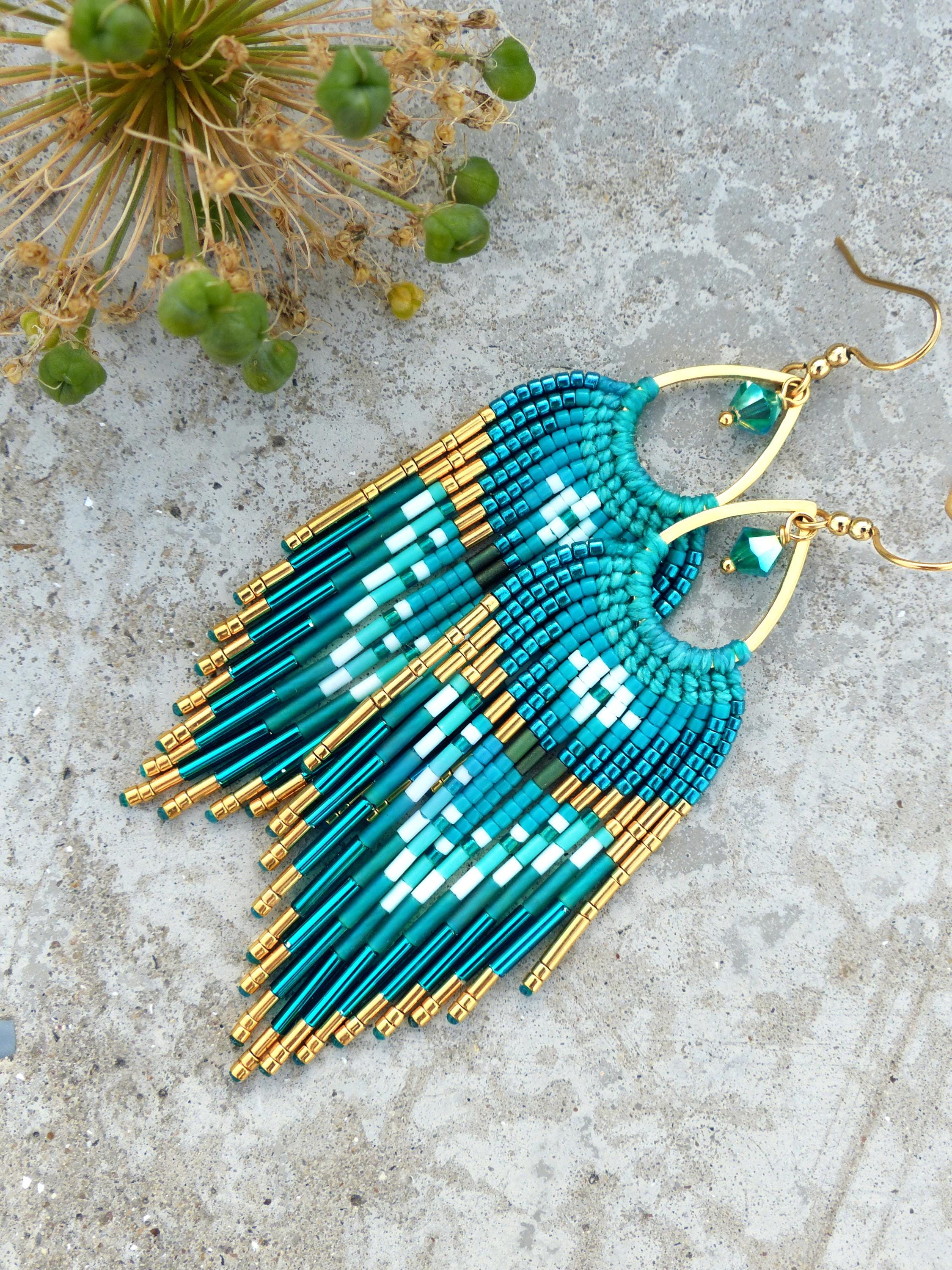 Peut inclure: Boucles d'oreilles en perles de couleur teal et or, avec un motif à franges. Les boucles d'oreilles présentent un motif géométrique blanc et teal, avec des accents dorés et des crochets d'oreilles. Mesurent environ 7.6 cm de long.
