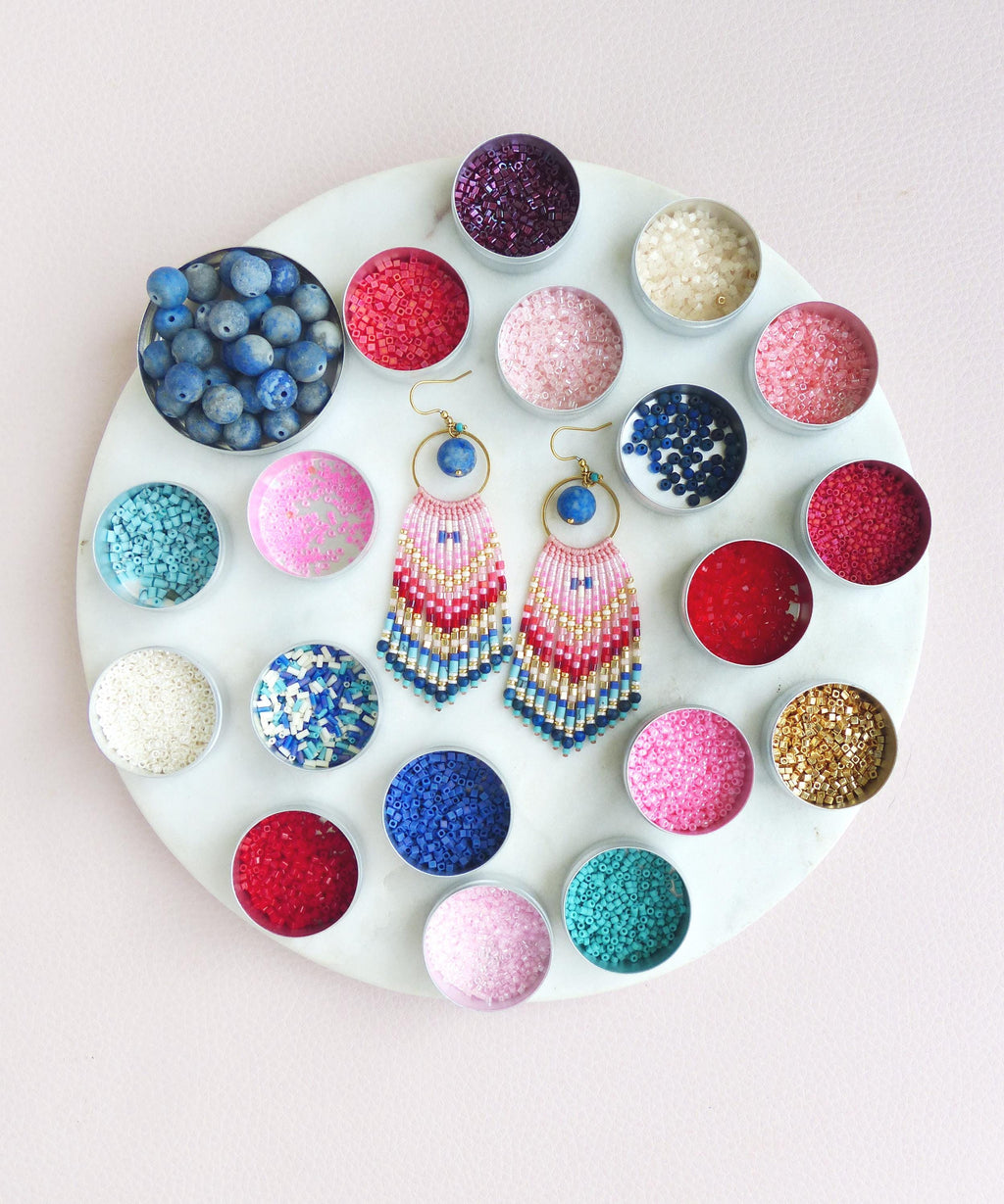 Peut inclure: Une paire de boucles d'oreilles à franges perlées avec un anneau doré, une perle bleue et un motif dégradé de perles de rocaille roses, rouges, dorées et bleues. Elles sont entourées de nombreux petits contenants métalliques remplis de perles.
