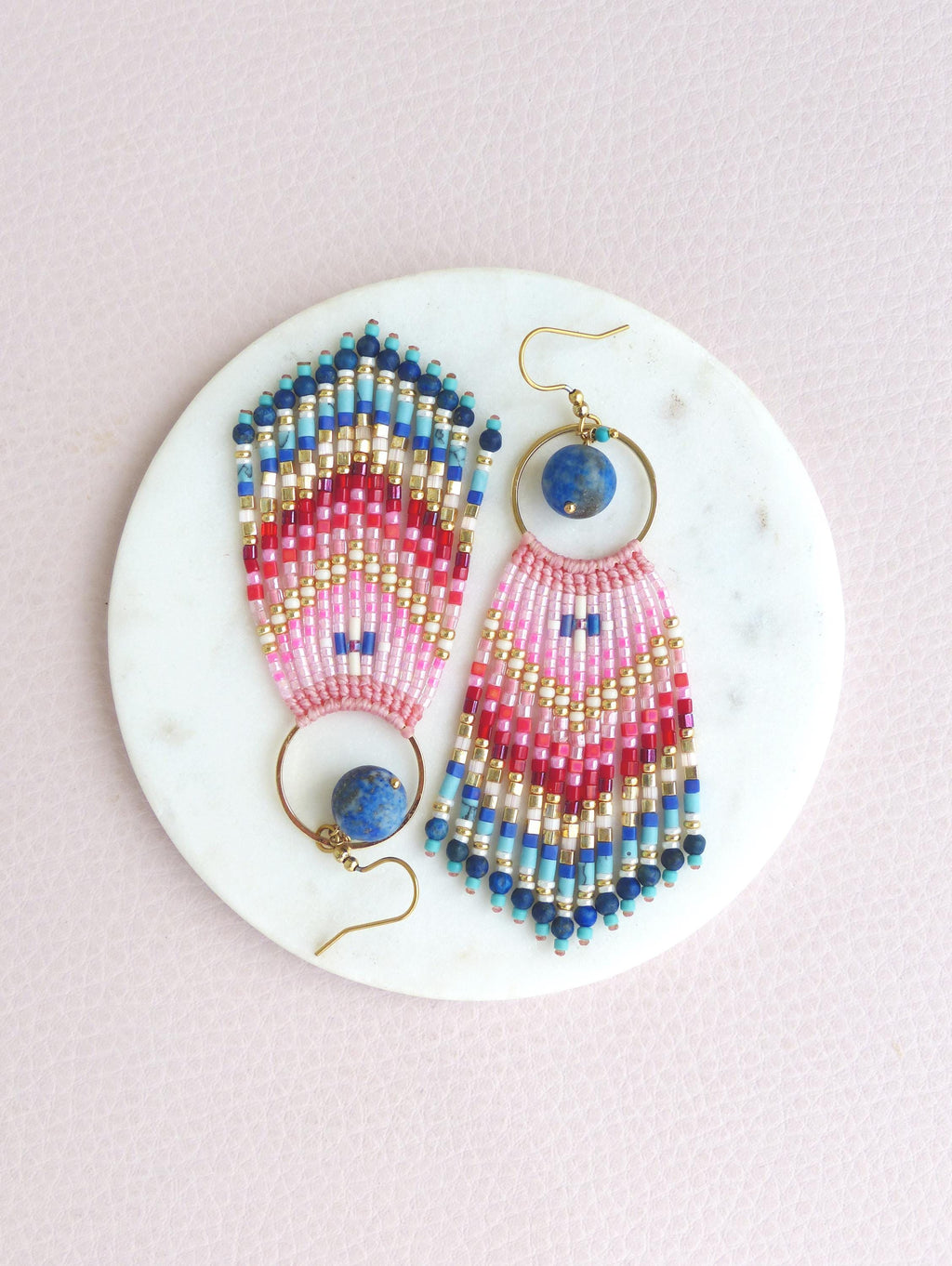 Peut inclure: Une paire de boucles d'oreilles pendantes avec un anneau doré, une pierre bleue ronde et une frange de perles multicolores. La frange présente un motif en chevron dans des tons de perles en verre rose, rouge, blanc, doré, bleu clair et bleu foncé.