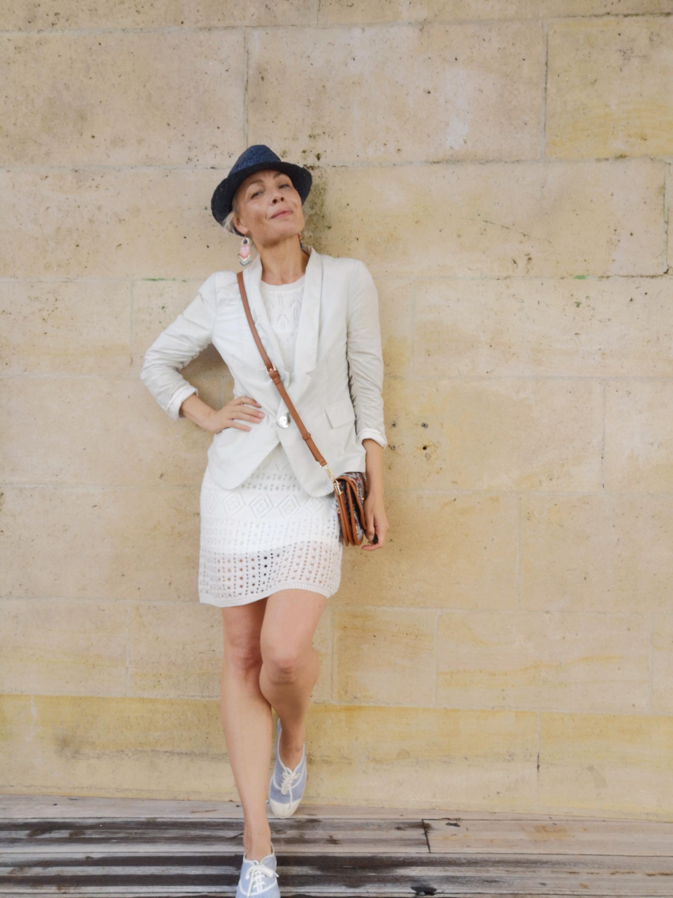 Peut inclure: Un chapeau fedora bleu marine, un blazer beige clair à manches longues, une robe en maille crochet blanche, un sac bandoulière en cuir marron avec une fine sangle, et des baskets en toile bleu clair à lacets blancs et semelles en caoutchouc.