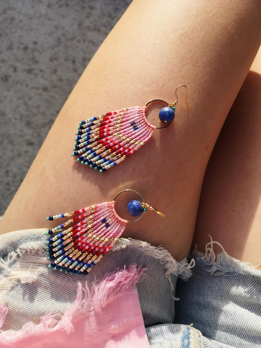 Peut inclure: Une paire de boucles d'oreilles pendantes avec un anneau doré attaché à une perle bleue ronde. Sous l'anneau, une frange en éventail composée de petites perles en verre dans un dégradé de rose, rouge, blanc, bleu clair et bleu foncé.