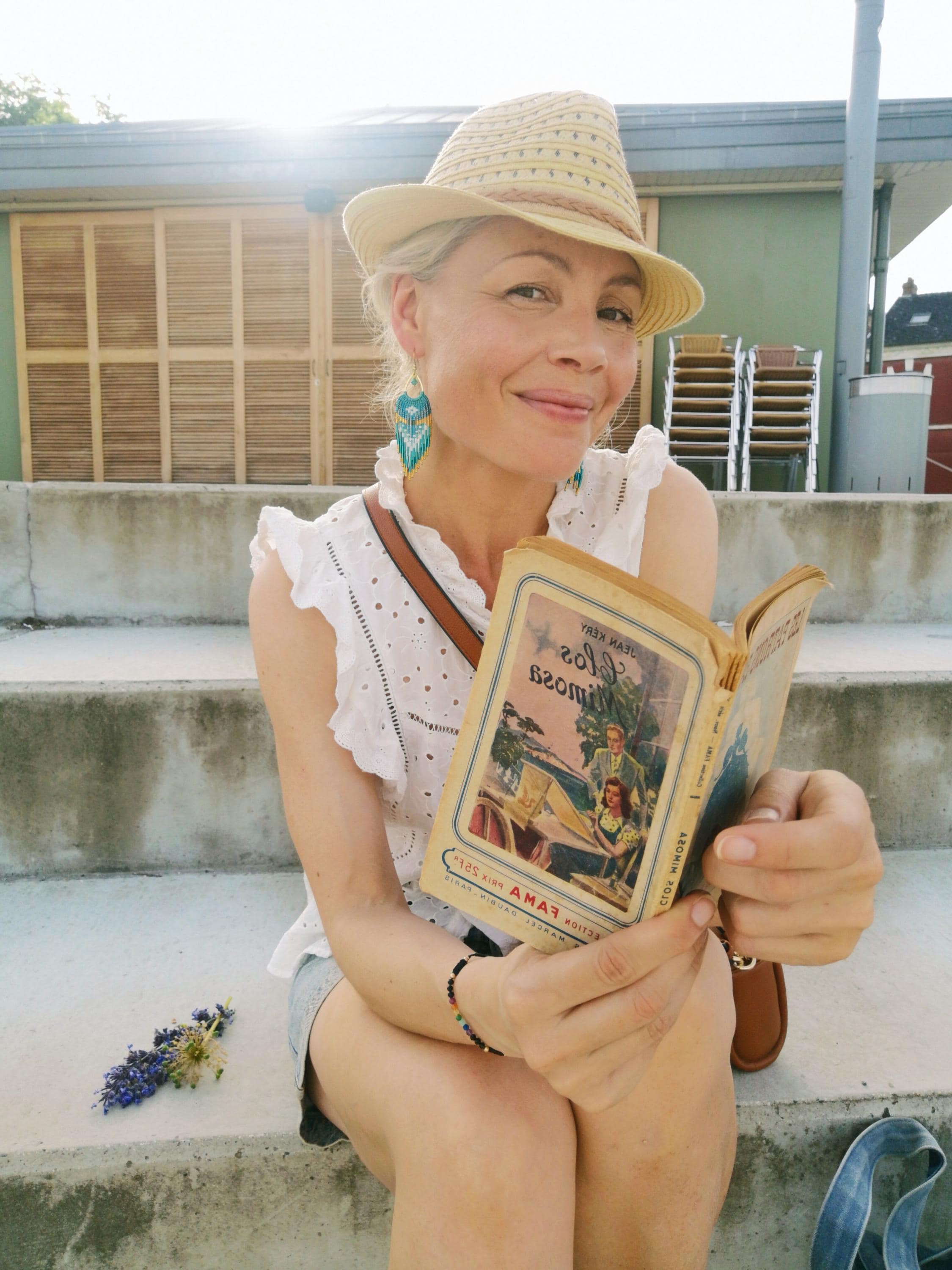 Peut inclure: Une femme portant un chapeau de paille et un haut blanc à œillets, tenant un livre vintage ouvert avec une couverture colorée. Elle est assise sur des marches en béton, avec une brindille de fleurs violettes à proximité. Le titre du livre est visible.