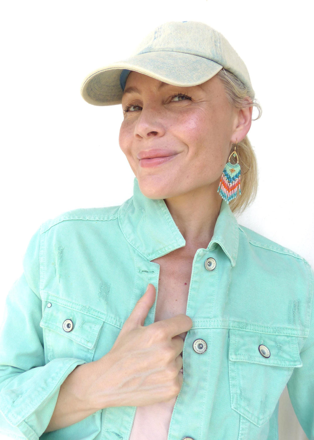 Peut inclure: Une veste en jean vert menthe clair avec des boutons en métal, une casquette de baseball en jean délavé clair et des boucles d'oreilles pendantes en perles avec un motif chevron turquoise, orange, blanc et doré, suspendues à un crochet doré.