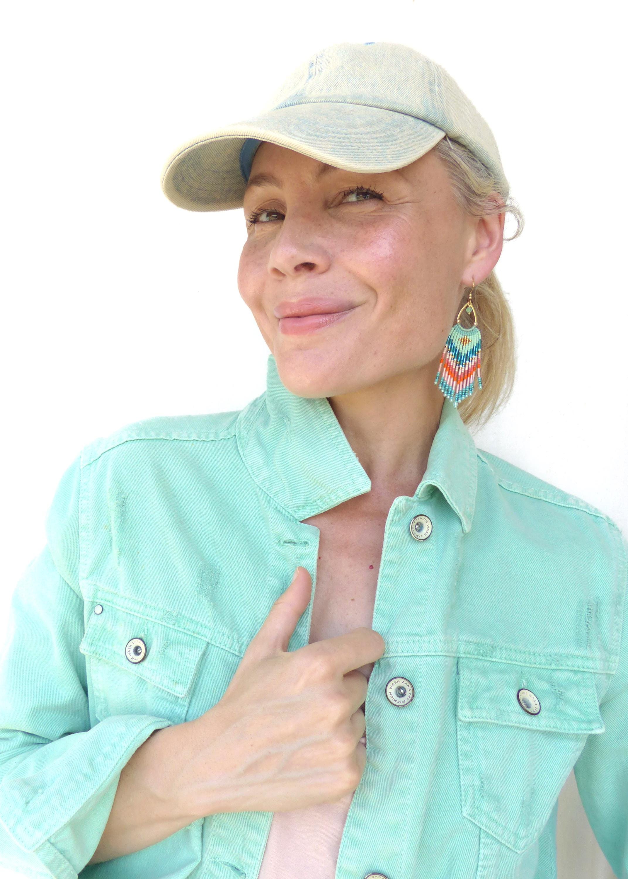 Peut inclure: Une veste en jean vert menthe clair avec des boutons en métal, une casquette de baseball en jean délavé clair et des boucles d'oreilles pendantes en perles avec un motif chevron turquoise, orange, blanc et doré, suspendues à un crochet doré.