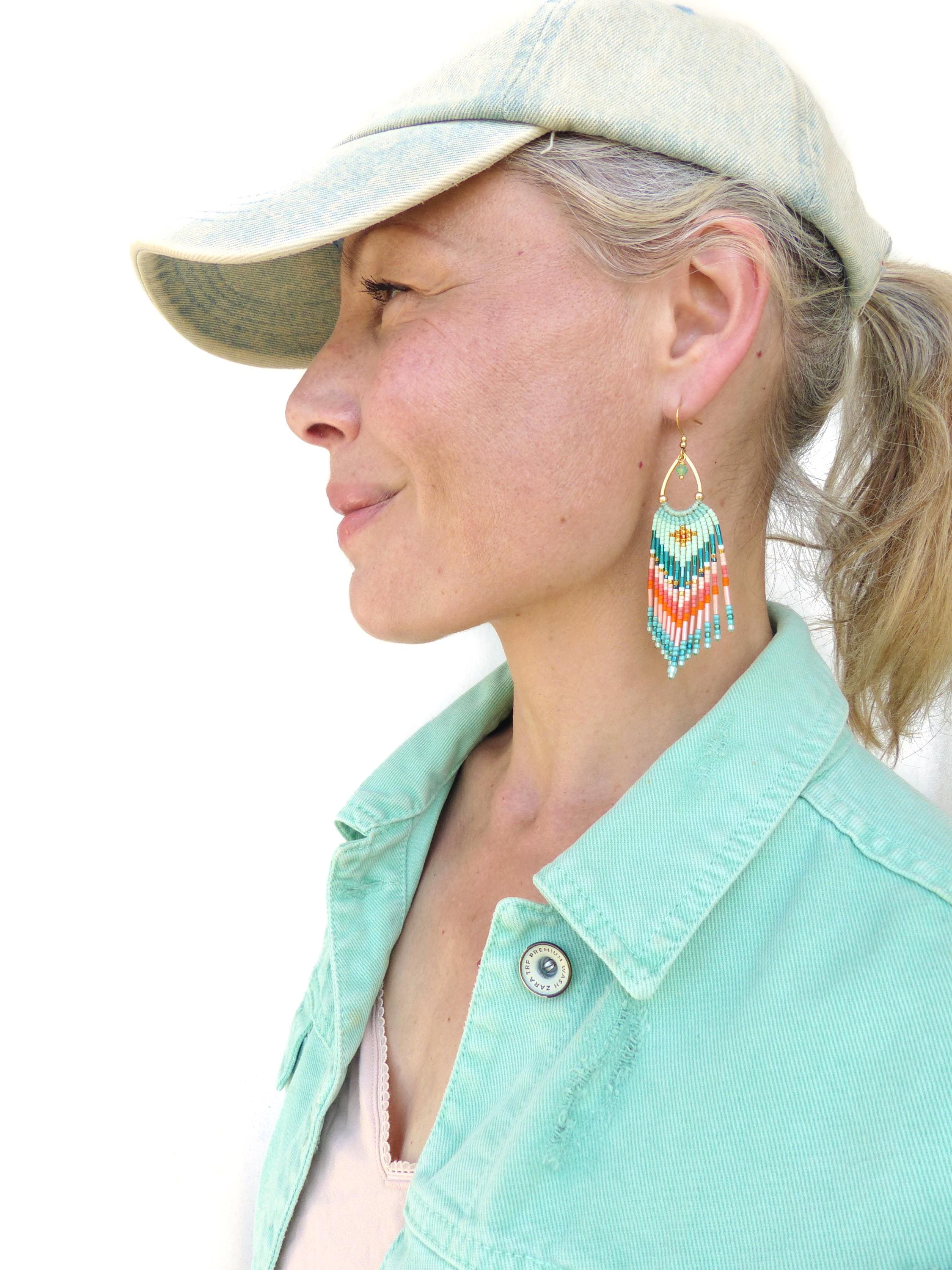 Peut inclure: Une paire de boucles d'oreilles à franges en perles en forme de goutte d'eau avec un motif géométrique dans des tons turquoise, blanc et orange. Les boucles d'oreilles sont suspendues à des crochets en métal doré et composées de perles en verre.