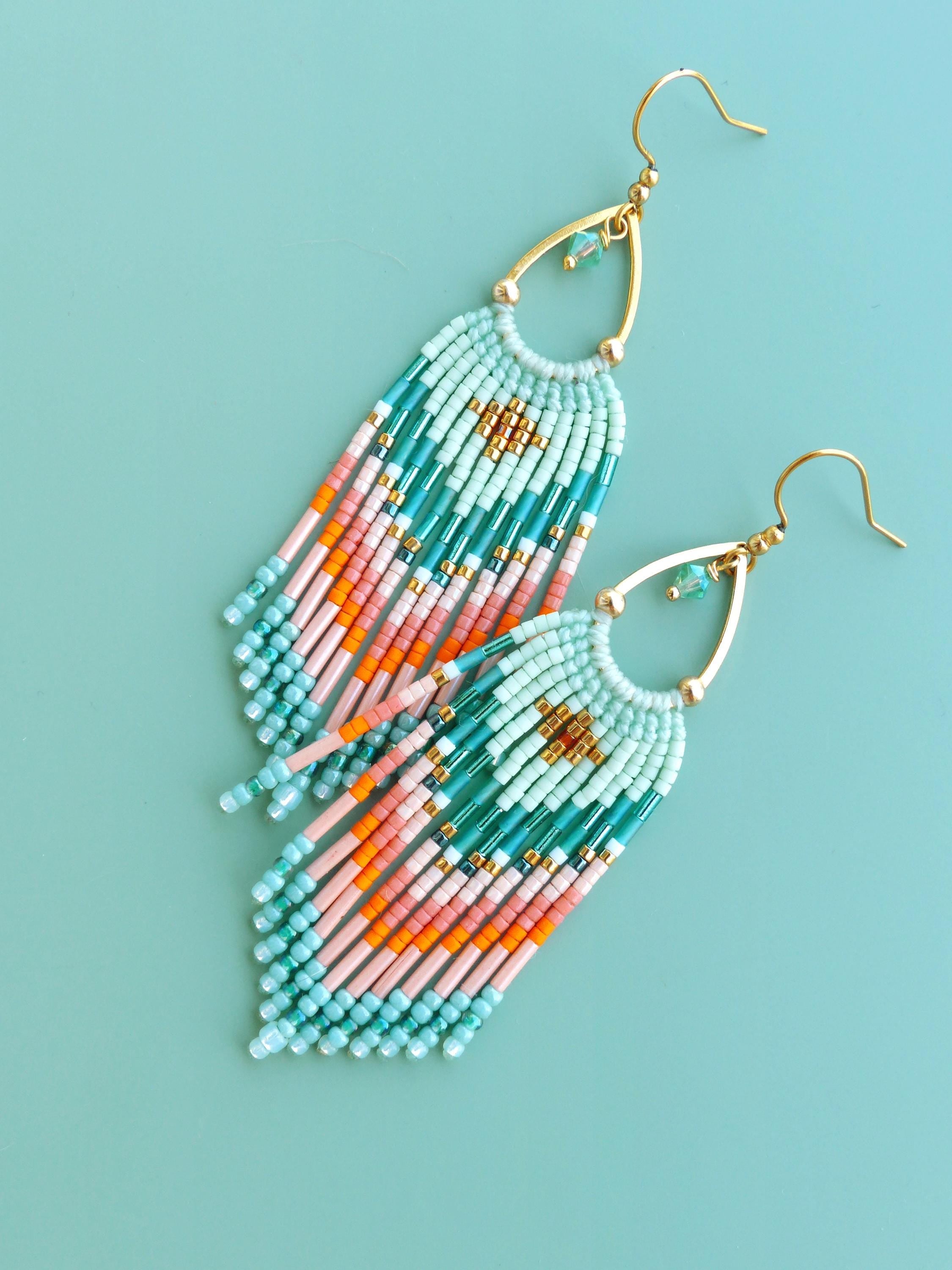 Peut inclure: Une paire de boucles d'oreilles pendantes avec des crochets dorés et des cadres en forme de goutte. Elles sont ornées de rangées de petites perles en verre dans des tons vert menthe, rose pâle et orange vif, formant un motif géométrique.