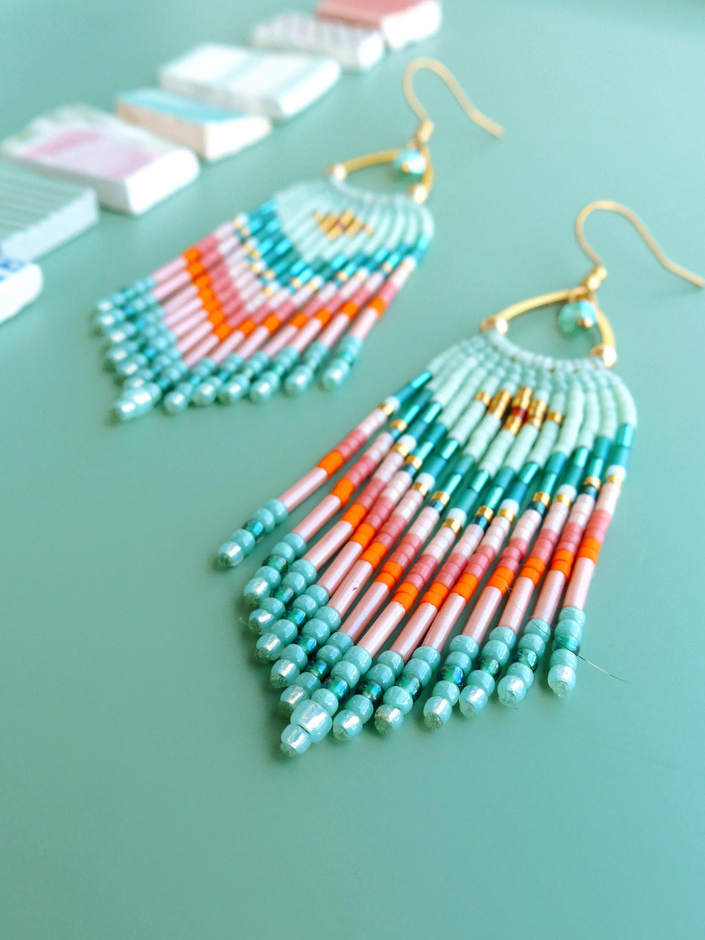 Peut inclure: Une paire de boucles d'oreilles pendantes composées de perles de rocaille en verre disposées en éventail dans des tons vert menthe, corail, orange et doré. Elles sont suspendues à des crochets en métal doré avec une perle au sommet.