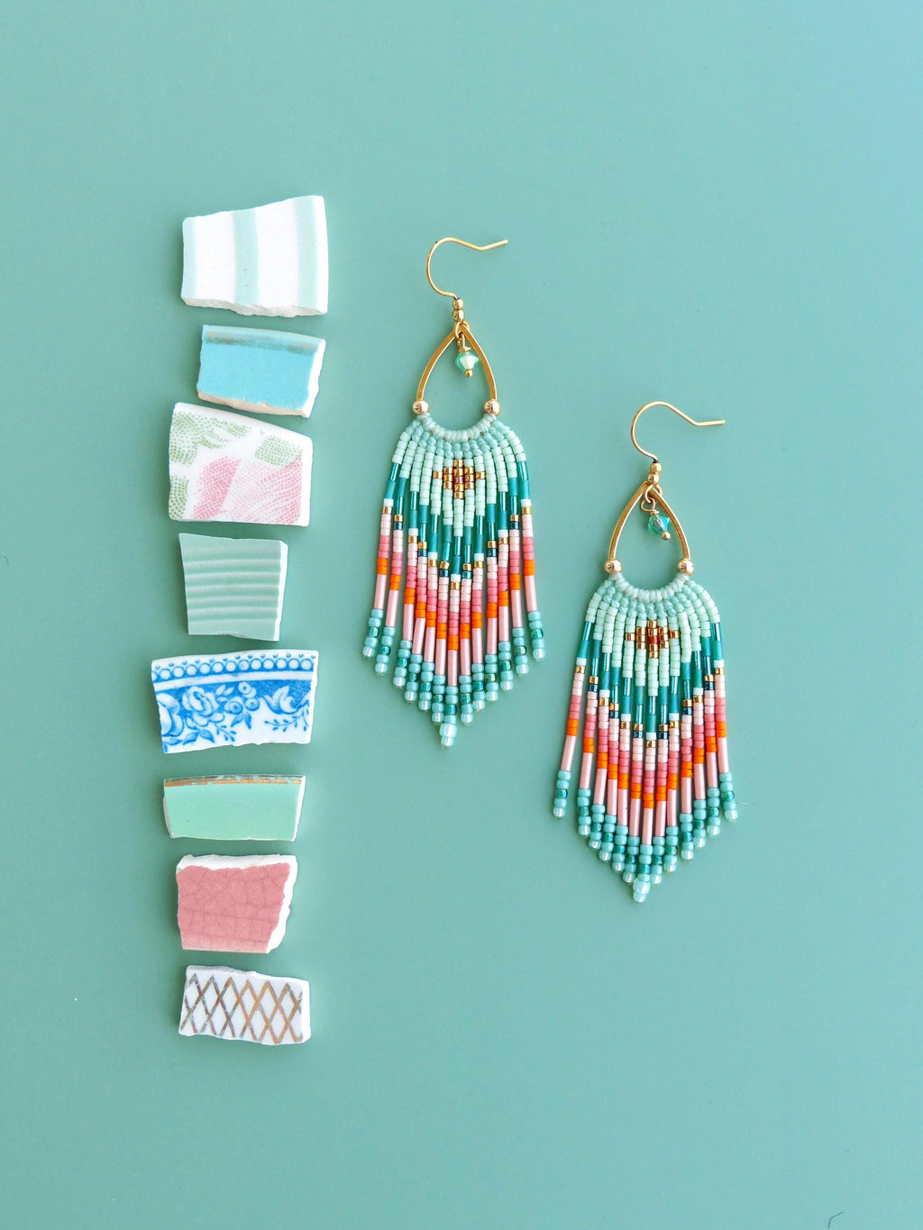 Peut inclure: Une paire de boucles d'oreilles à franges en perles en forme de goutte, arborant un motif géométrique dans des tons turquoise, corail, rose et doré, suspendues à des crochets dorés. Huit éclats de céramique assortis sont disposés à côté.
