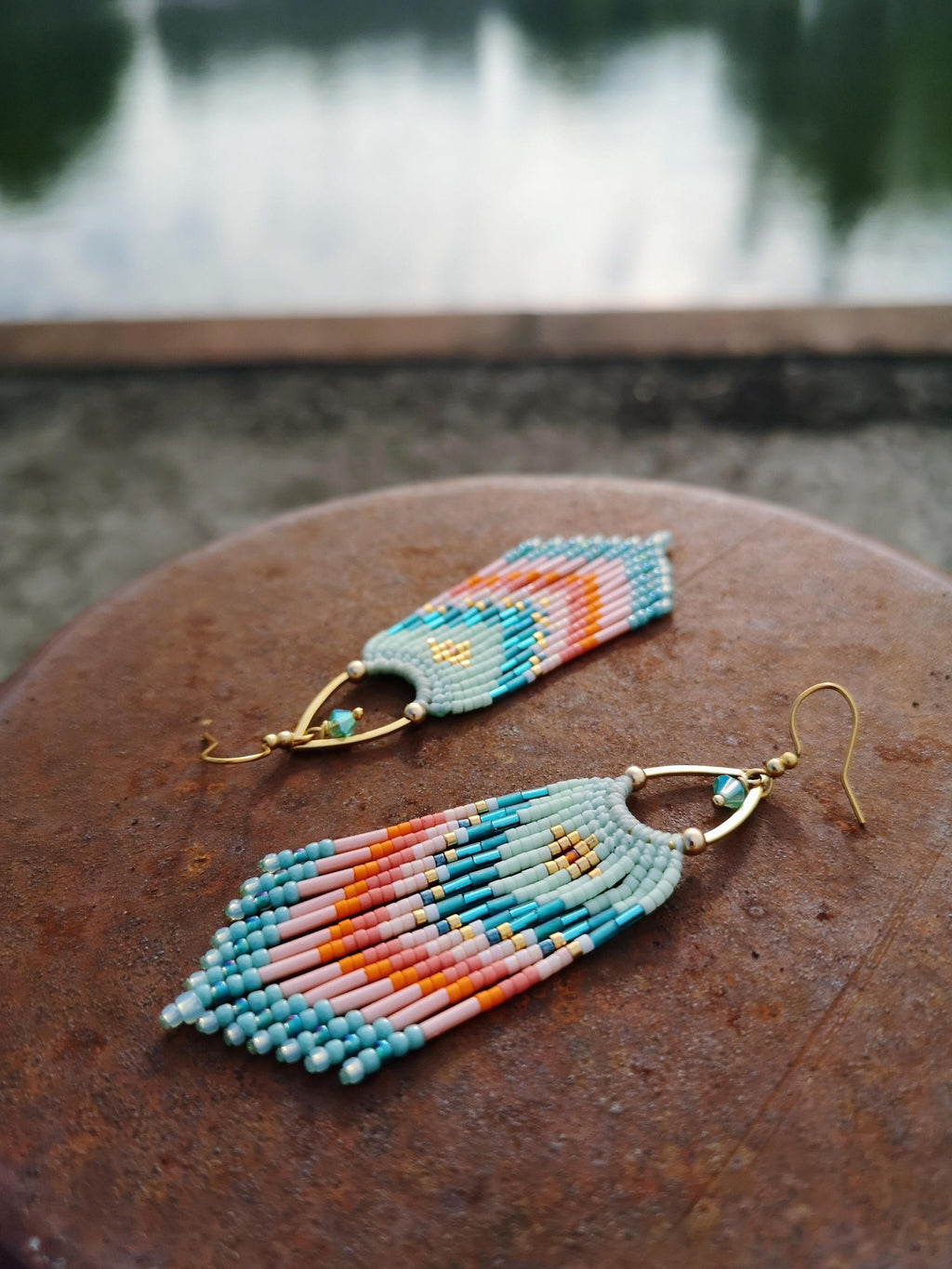 Peut inclure: Une paire de boucles d'oreilles à franges composées de petites perles en verre dans des tons bleu clair, pêche, orange et doré. Les boucles présentent une monture triangulaire en métal doré avec une perle turquoise et des crochets dorés.