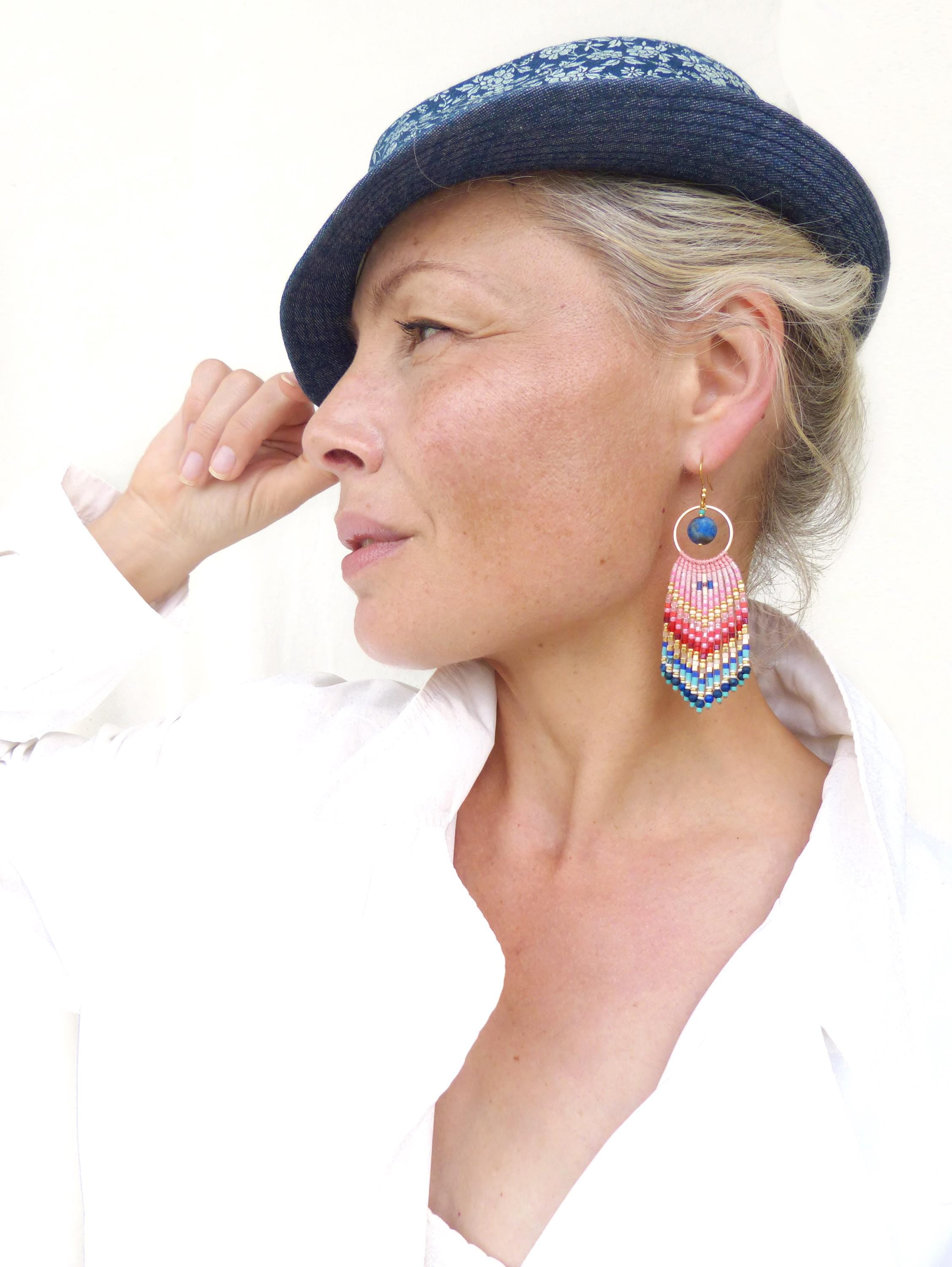Peut inclure: Une boucle d'oreille pendante composée d'un anneau argenté avec une perle bleue, relié à une frange triangulaire en perles de verre tissées dégradées rose, rouge, blanc et bleu, suspendue à un crochet doré.
