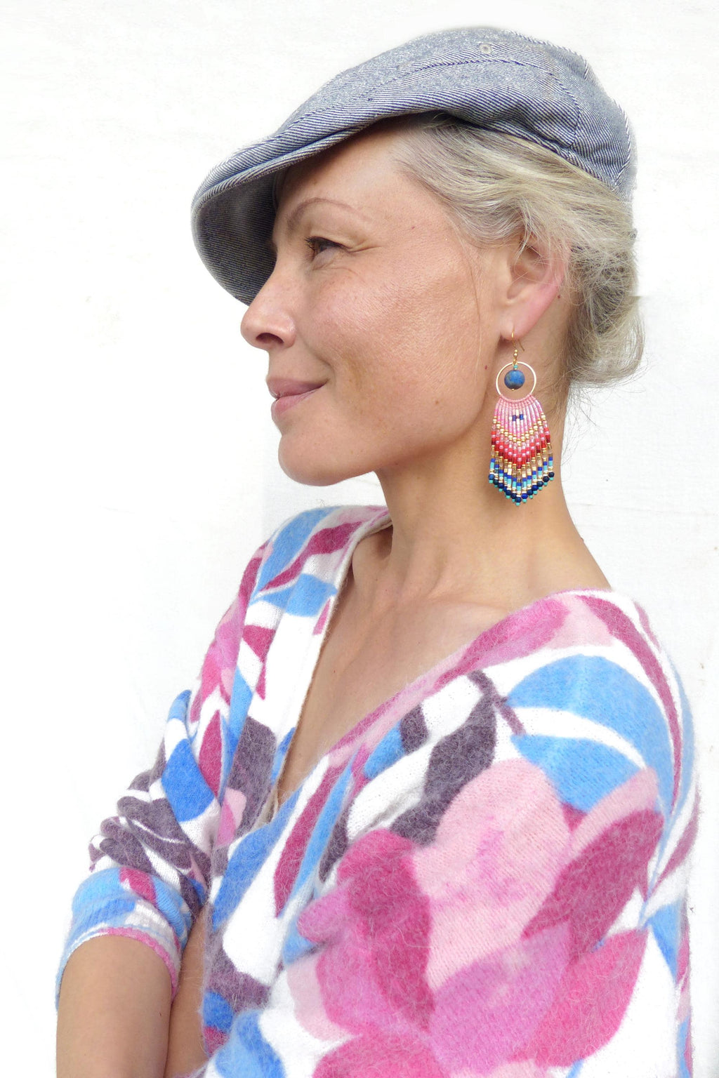 Peut inclure: Une paire de boucles d'oreilles pendantes avec un anneau doré, une perle bleue unique et une frange de perles multicolores dans des tons rose, rouge, jaune et bleu. Les boucles sont portées avec une casquette grise et un pull blanc à motif floral.