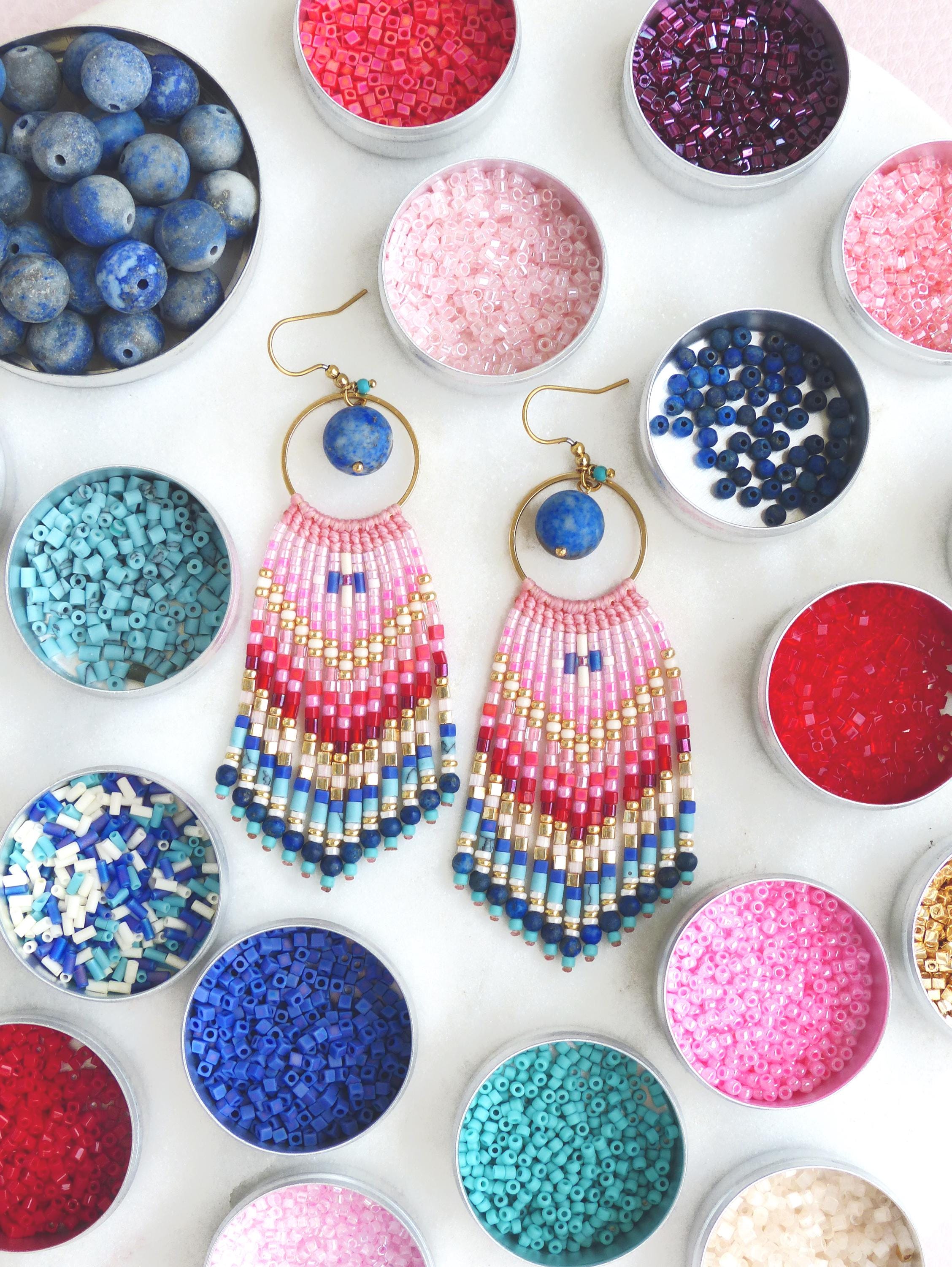 Peut inclure: Une paire de boucles d'oreilles pendantes avec des anneaux dorés, des perles en pierre bleue et des franges de perles de rocaille multicolores dans des tons de rose, rouge, or et bleu. Elles sont entourées de boîtes métalliques remplies de perles.