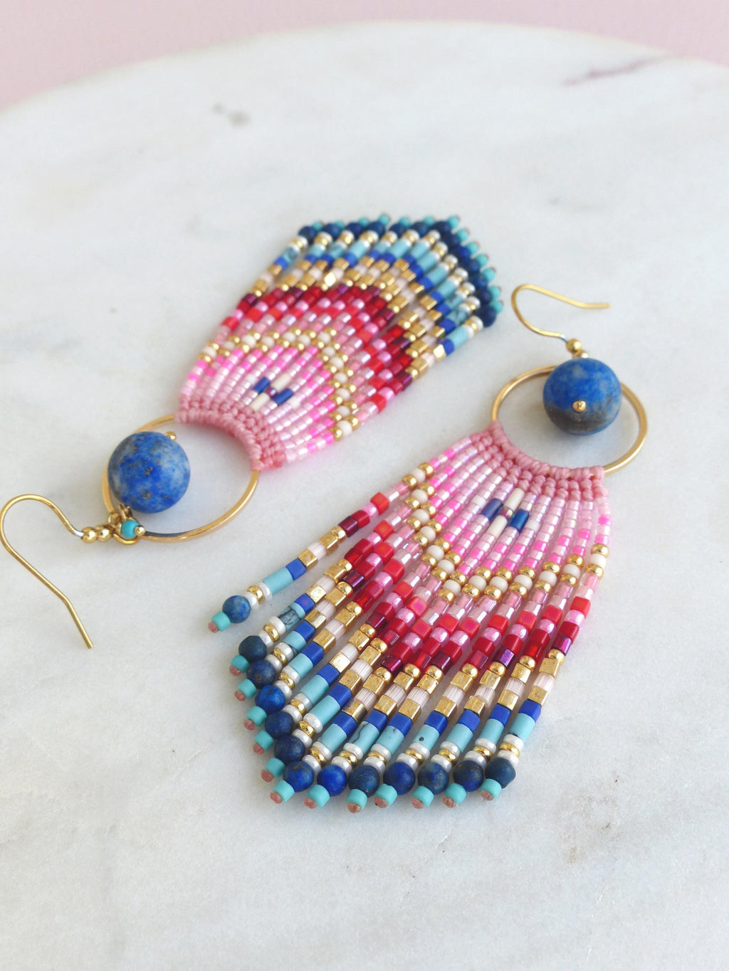 Peut inclure: Une paire de boucles d'oreilles pendantes avec un crochet doré et un anneau circulaire retenant une perle ronde en lapis-lazuli bleu. Sous l'anneau, une section en éventail de perles de rocaille tissées en rose, rouge, blanc, or et bleu.