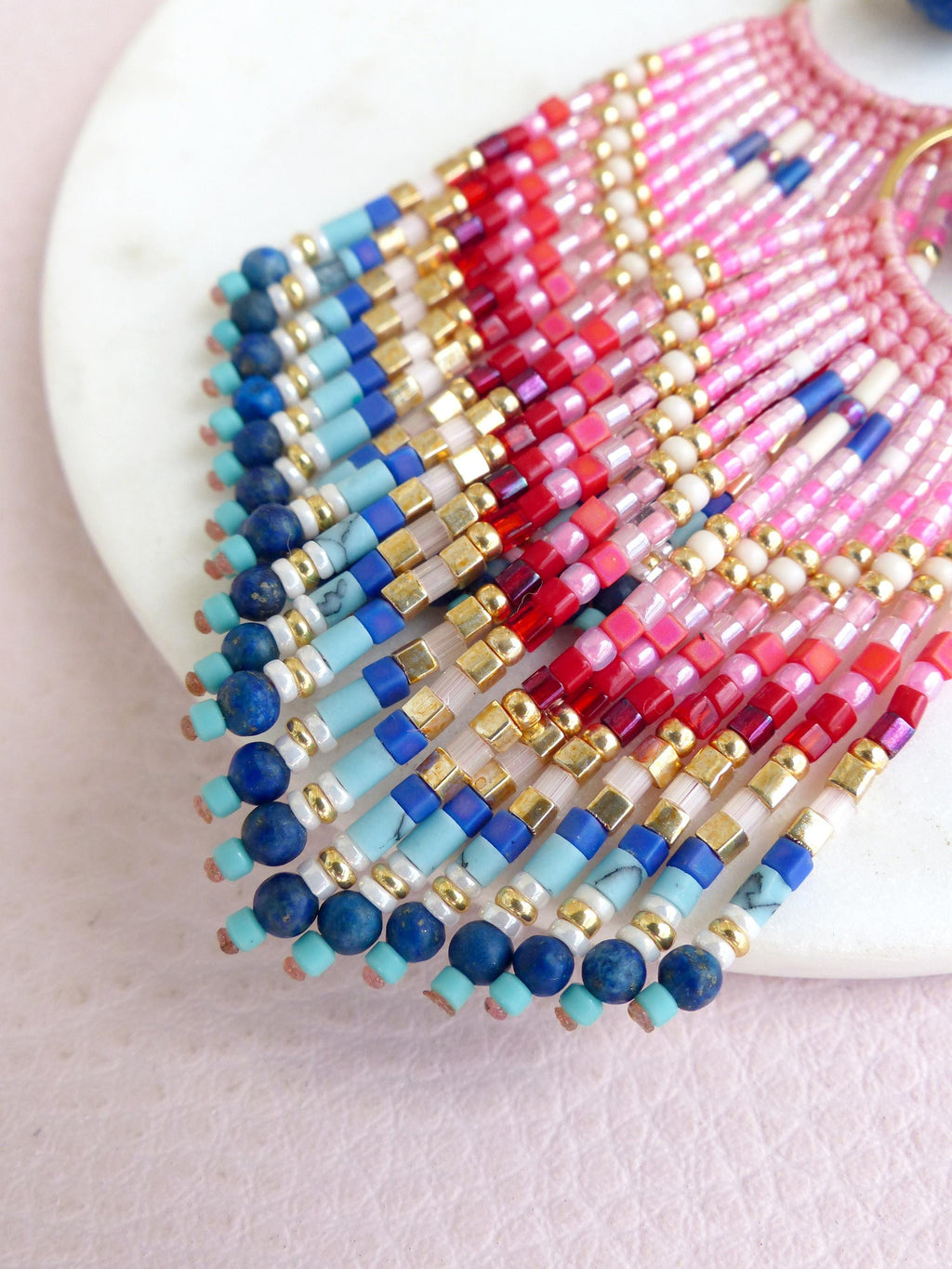 Peut inclure: Une paire de boucles d'oreilles en éventail composées de perles de rocaille multicolores dans des tons rose, rouge, bleu et doré. Le design présente une frange de perles pendantes, incluant des pierres rondes bleu foncé, turquoise et dorées.