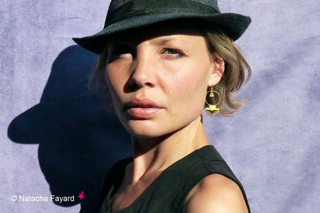 Peut inclure: Une femme portant un chapeau fedora noir et un haut noir sans manches avec une paire de boucles d'oreilles créoles dorées avec un pompon jaune et noir.