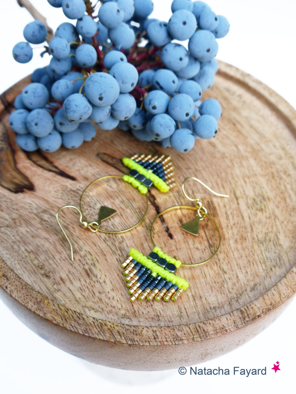 Peut inclure: Une paire de boucles d'oreilles créoles dorées avec un motif géométrique. Les boucles d'oreilles sont faites de perles noires et vert fluo. Les boucles d'oreilles sont sur une surface en bois.