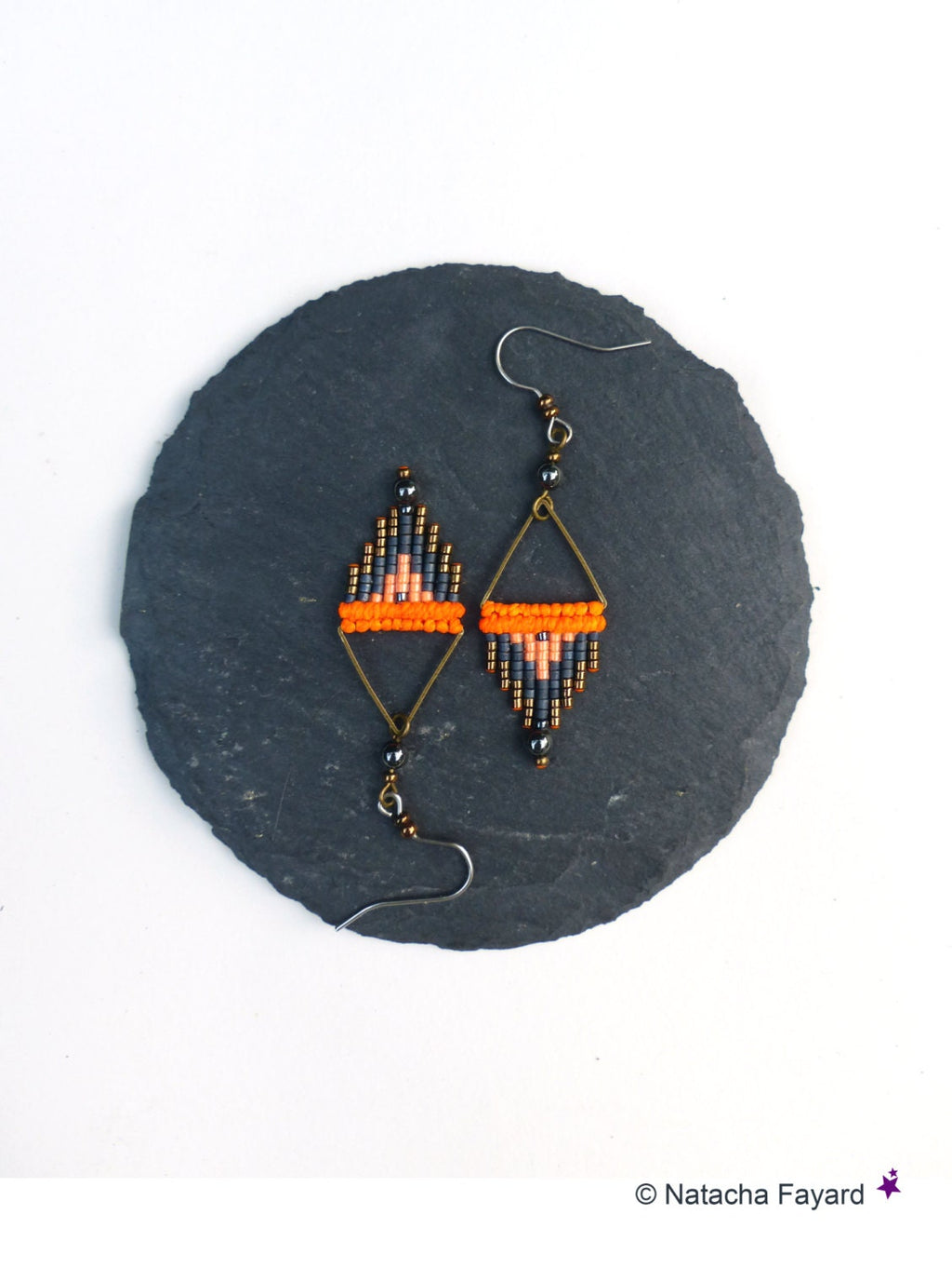 Peut inclure: Une paire de boucles d'oreilles en perles orange, grises et noires avec des accents de fil doré. Les boucles d'oreilles sont suspendues à une surface de pierre gris foncé.