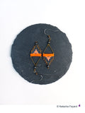 Peut inclure: Une paire de boucles d'oreilles en perles orange, grises et noires avec des accents de fil doré. Les boucles d'oreilles sont suspendues à une surface de pierre gris foncé.