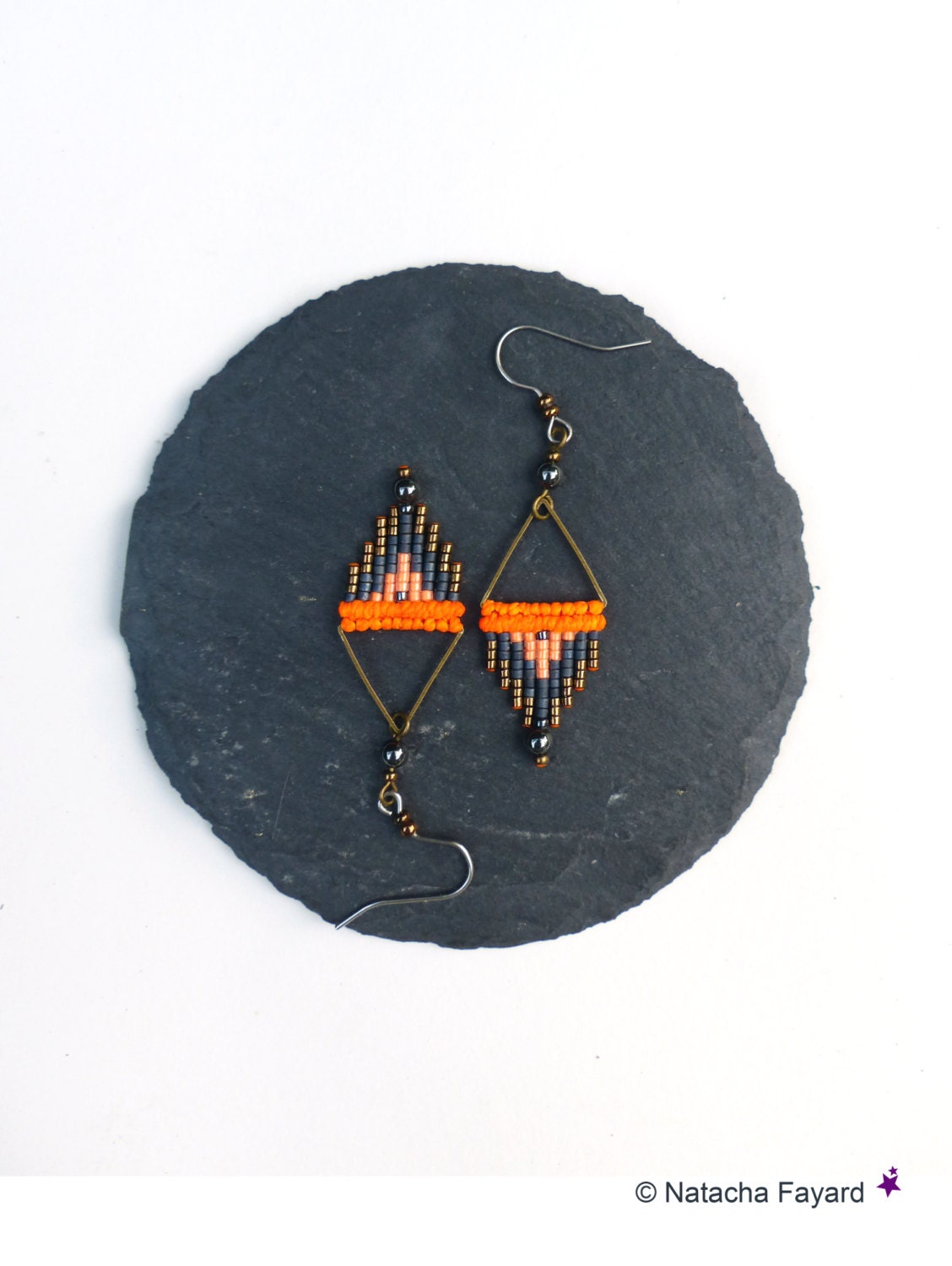 Peut inclure: Une paire de boucles d'oreilles en perles orange, grises et noires avec des accents de fil doré. Les boucles d'oreilles sont suspendues à une surface de pierre gris foncé.