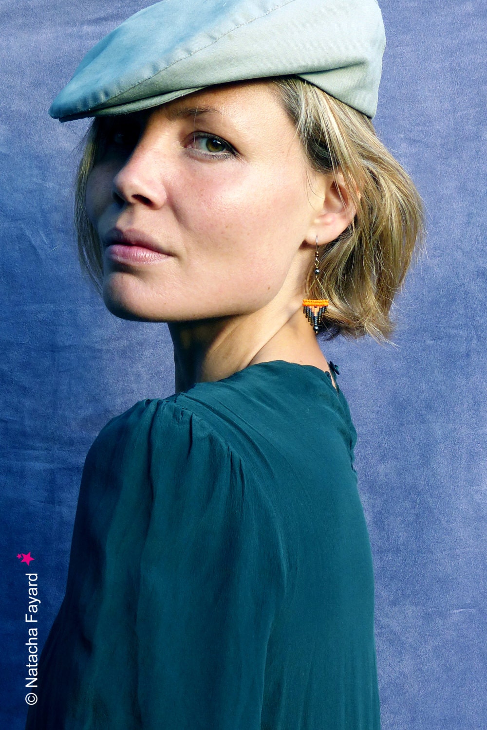 Peut inclure: Une femme aux cheveux blonds porte une casquette plate grise clair et une chemise vert sarcelle. Elle porte une paire de petites boucles d'oreilles pendantes avec des perles orange et noires.