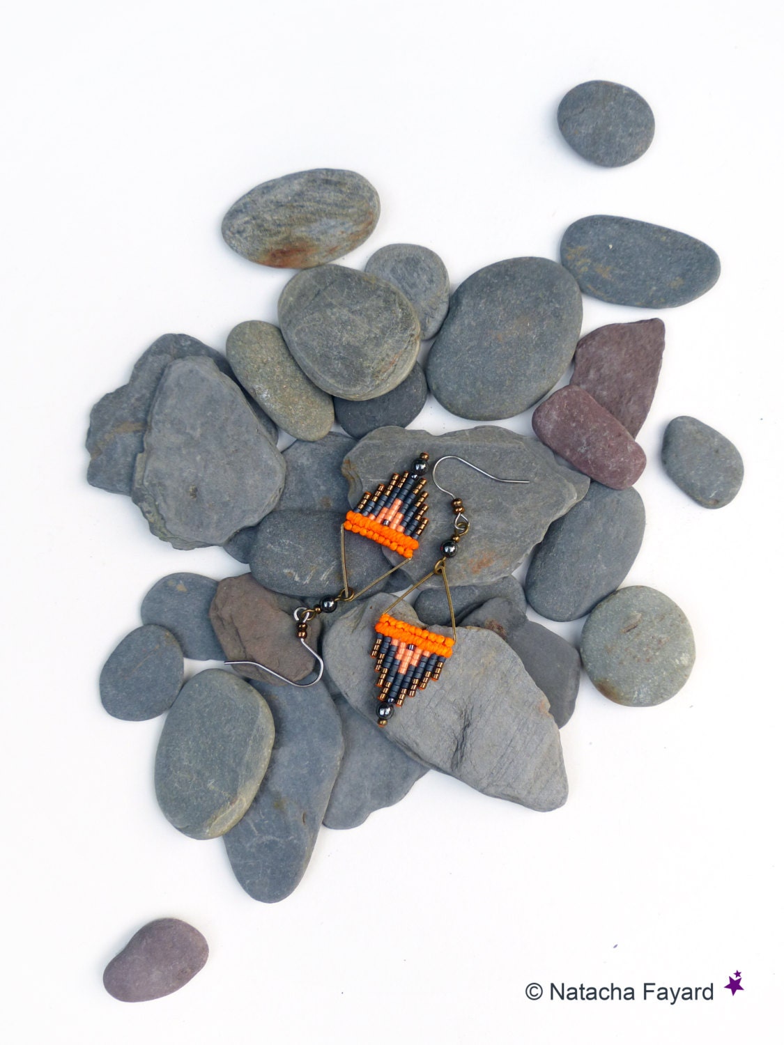 Peut inclure: Une paire de boucles d'oreilles en perles orange et noires avec un motif géométrique, disposées sur un lit de pierres grises.