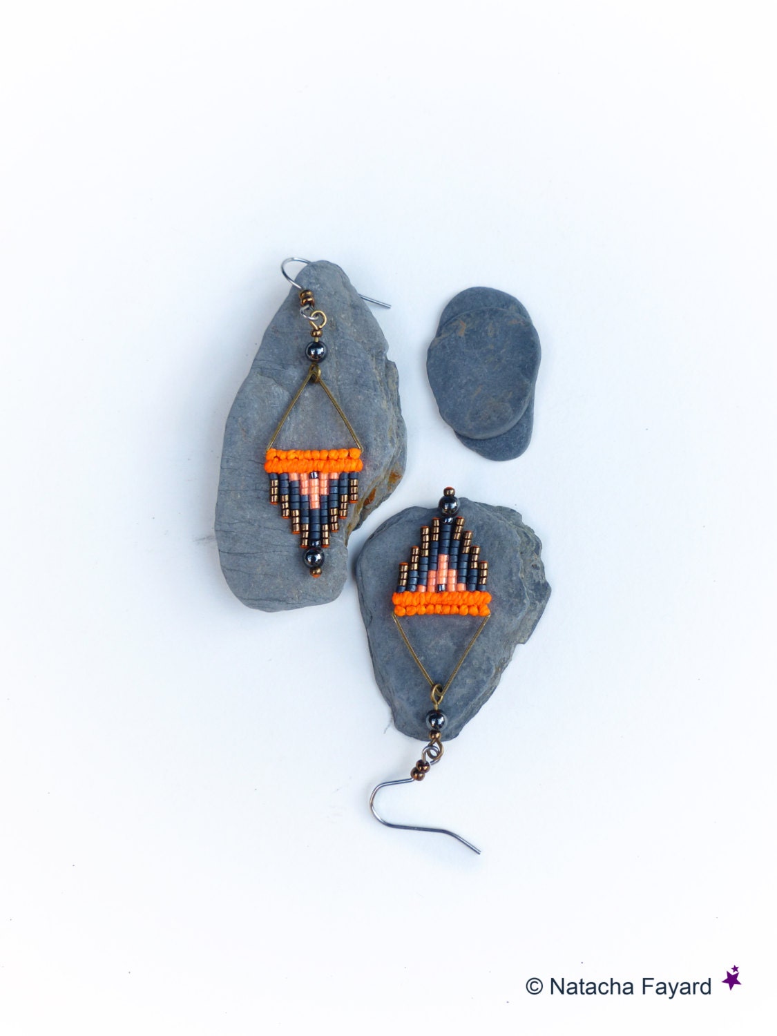 Peut inclure: Une paire de boucles d'oreilles en perles orange, noires et cuivre avec un motif géométrique. Les boucles d'oreilles sont suspendues à des pierres grises.
