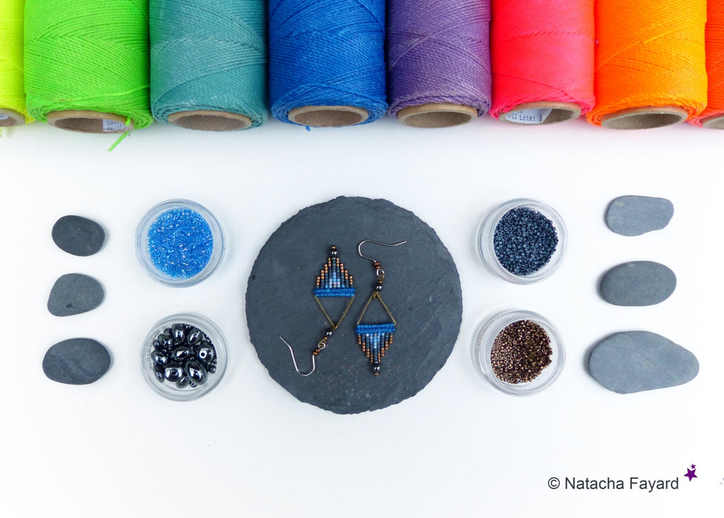 Peut inclure: Une paire de boucles d'oreilles en perles bleues et cuivre avec un motif géométrique, présentée sur une dalle de schiste noire avec diverses perles et pierres dans de petits récipients en verre.