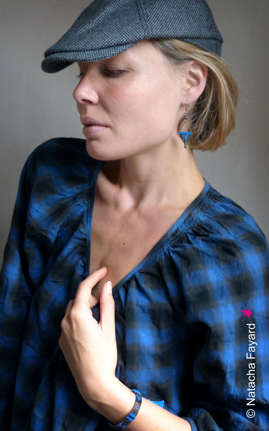 Peut inclure: Une femme portant une chemise à carreaux bleus et noirs avec une casquette de gavroche grise. Elle porte un bracelet bleu et noir en perles au poignet et une paire de boucles d'oreilles pendantes bleues et argentées.