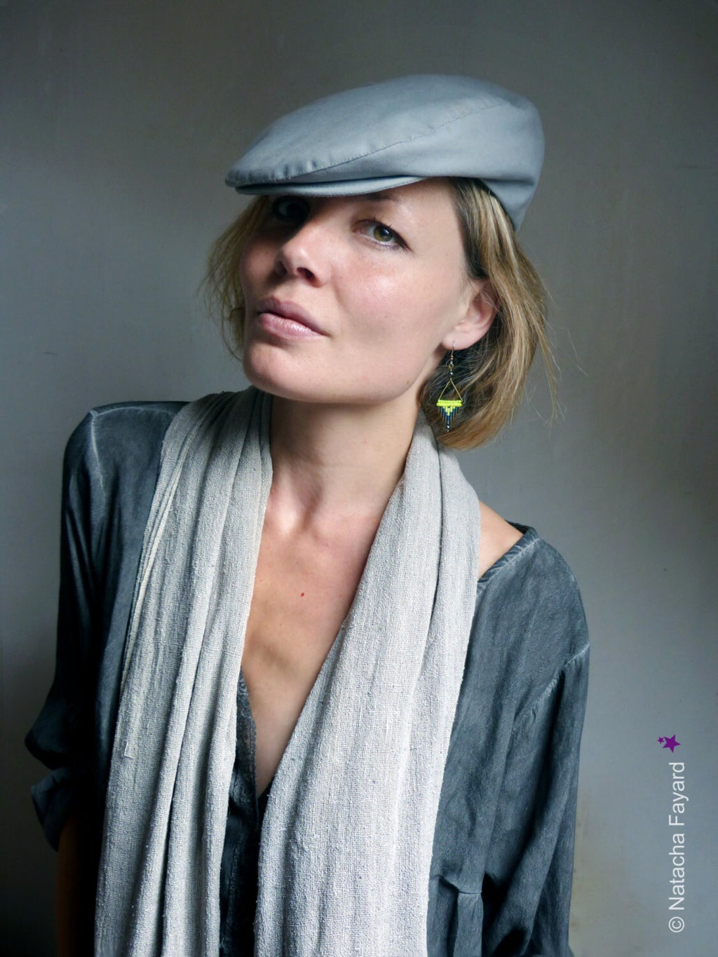 Peut inclure: Une femme portant une casquette plate grise, une écharpe grise et une chemise grise. Elle porte une paire de boucles d'oreilles jaunes et noires.