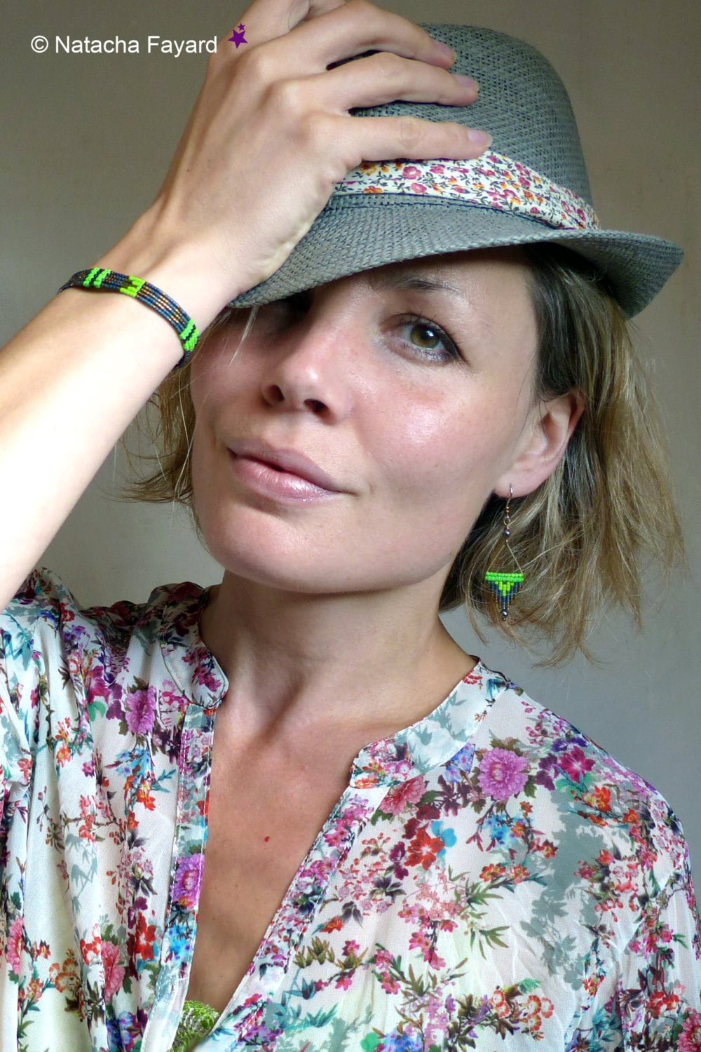 Peut inclure: Une femme portant un chapeau fedora gris avec un ruban floral, un chemisier à imprimé floral et un bracelet en perles noires et vertes. Elle porte également une paire de boucles d'oreilles en perles vertes et noires.