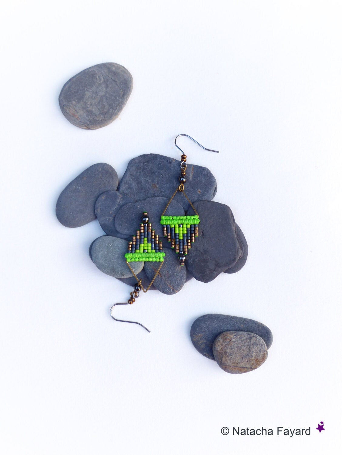 Peut inclure: Une paire de boucles d'oreilles en perles vertes et noires avec un motif géométrique. Les boucles d'oreilles sont suspendues à des crochets argentés et sont présentées sur un lit de pierres grises.