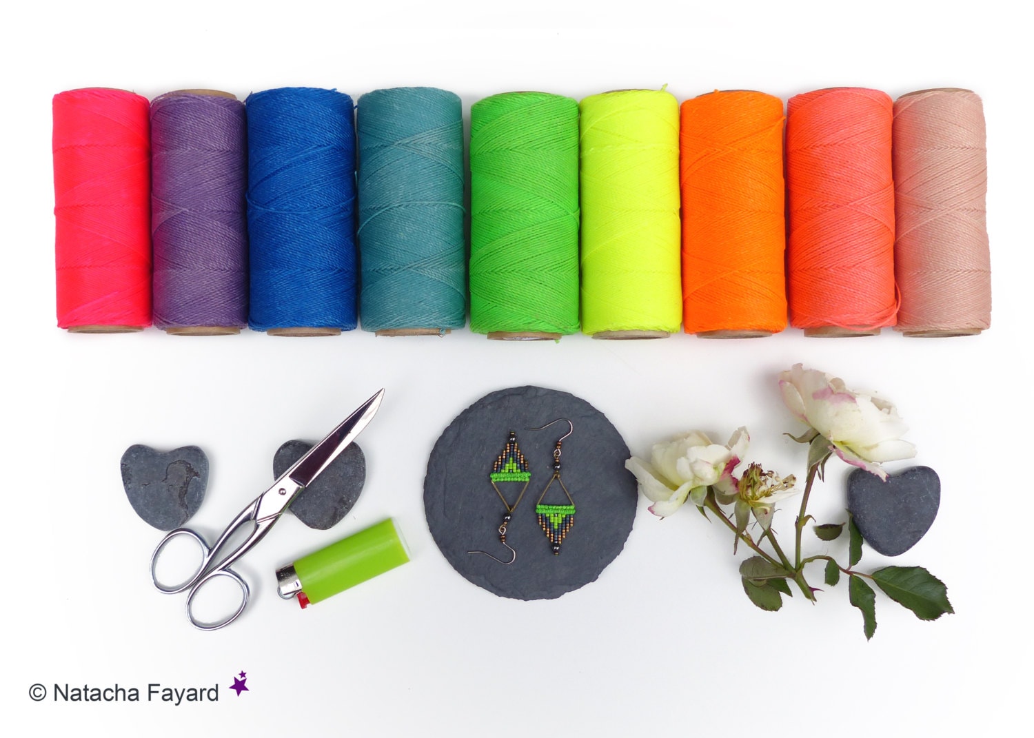 Peut inclure: Une vue de dessus de bobines de fil colorées, une paire de ciseaux, un briquet, deux pierres en forme de cœur, une dalle ardoise ronde avec une paire de boucles d'oreilles en perles vertes et brunes, et une brindille de roses blanches.