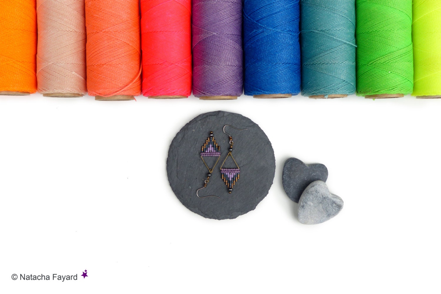 Peut inclure: Une paire de boucles d'oreilles en perles violettes et brunes avec un motif géométrique, présentée sur un cercle de schiste noir avec deux pierres en forme de cœur grises.