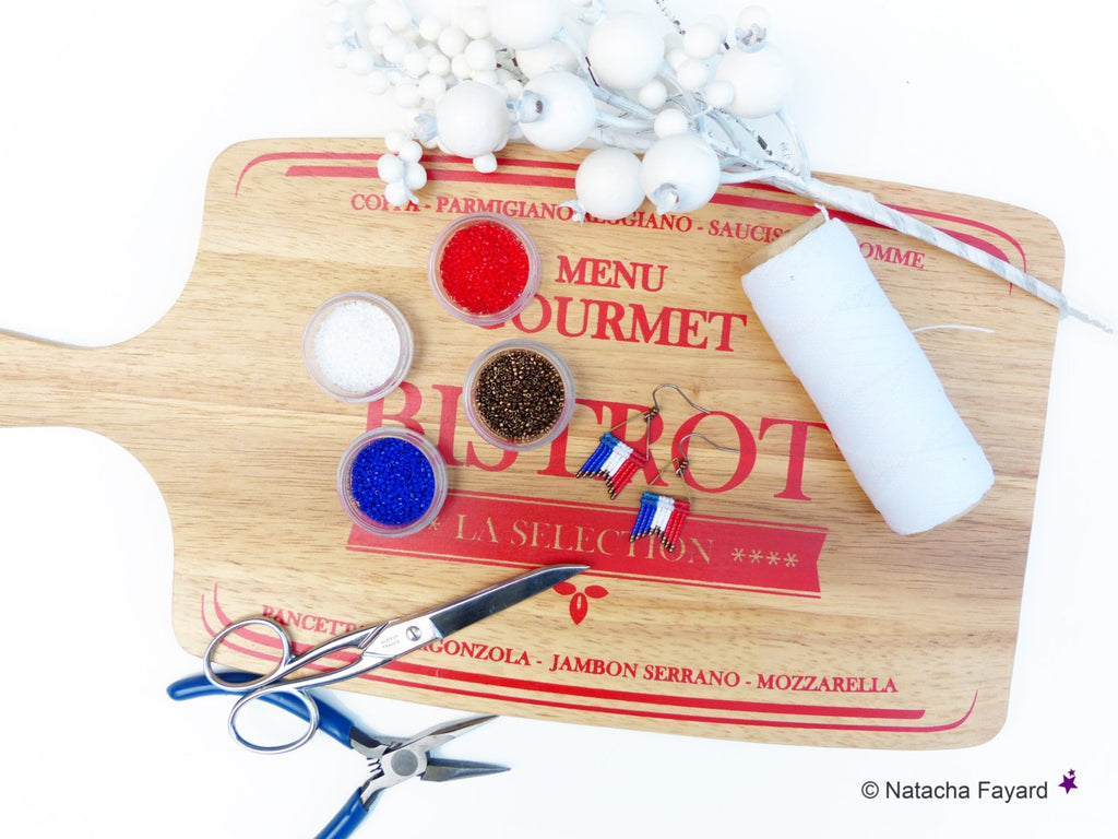Peut inclure: Une planche à découper en bois avec les mots "Bistrot" et "La Selection" imprimés dessus. Il y a quatre petits bols de perles rouges, blanches, bleues et brunes. Il y a aussi deux paires de boucles d'oreilles avec des perles rouges, blanches et bleues, une paire de ciseaux et une pince.