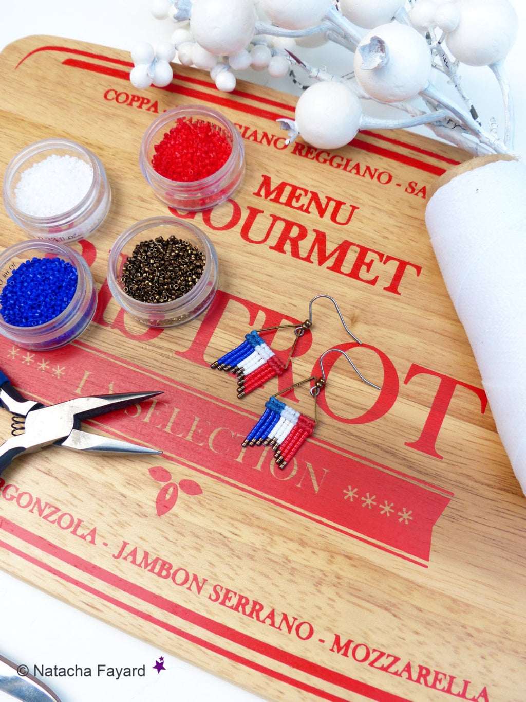 Peut inclure: Un plan rapproché d'une planche à découper en bois avec les mots "MENU GOURMET" imprimés en rouge. Il y a quatre petits contenants de perles en rouge, blanc, bleu et or. Une paire de boucles d'oreilles avec des perles rouges, blanches et bleues sont sur la planche. Les boucles d'oreilles sont en forme de drapeau français. Il y a aussi des pinces sur la planche.
