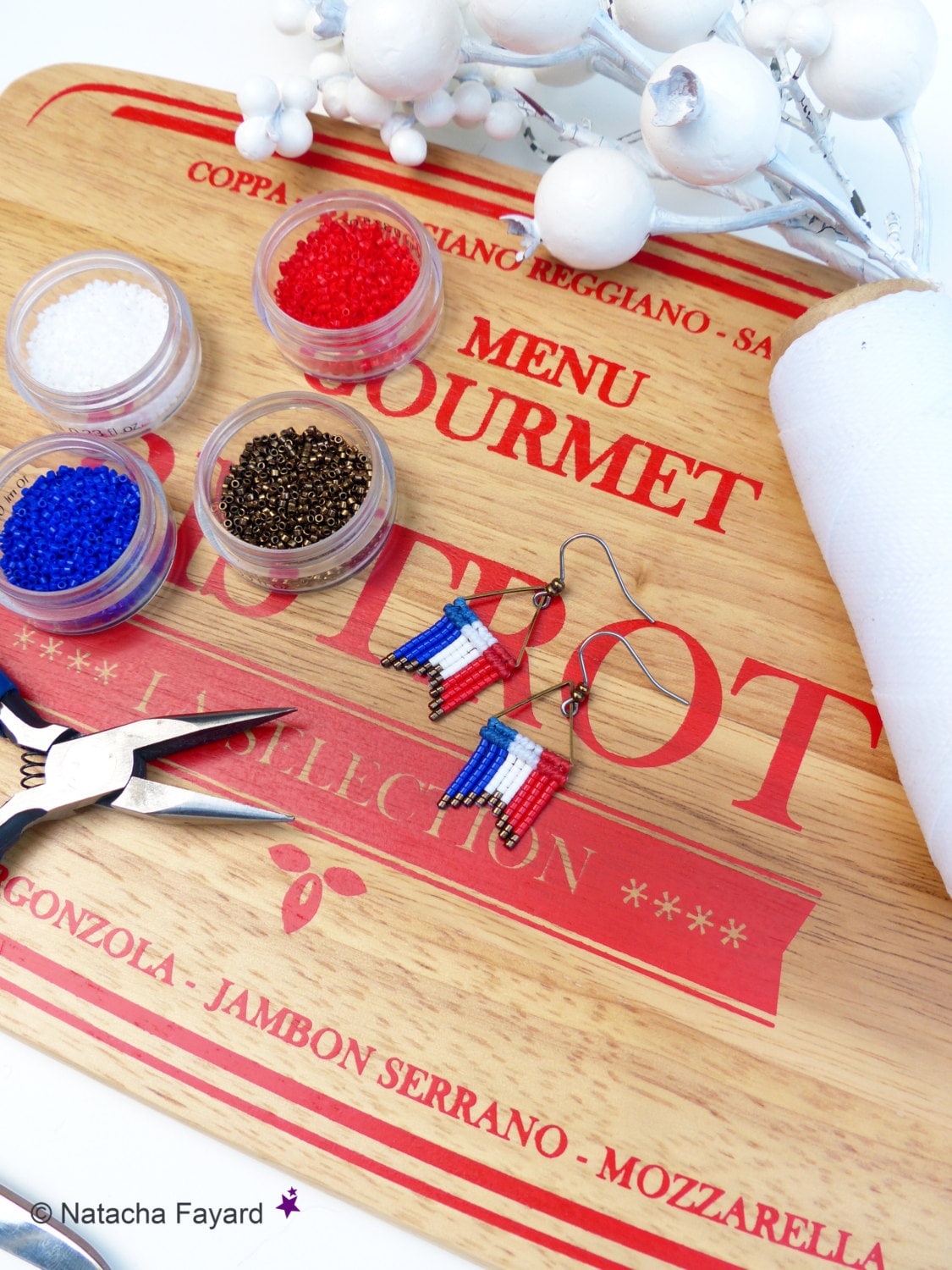 Peut inclure: Un plan rapproché d'une planche à découper en bois avec les mots "MENU GOURMET" imprimés en rouge. Il y a quatre petits contenants de perles en rouge, blanc, bleu et or. Une paire de boucles d'oreilles avec des perles rouges, blanches et bleues sont sur la planche. Les boucles d'oreilles sont en forme de drapeau français. Il y a aussi des pinces sur la planche.