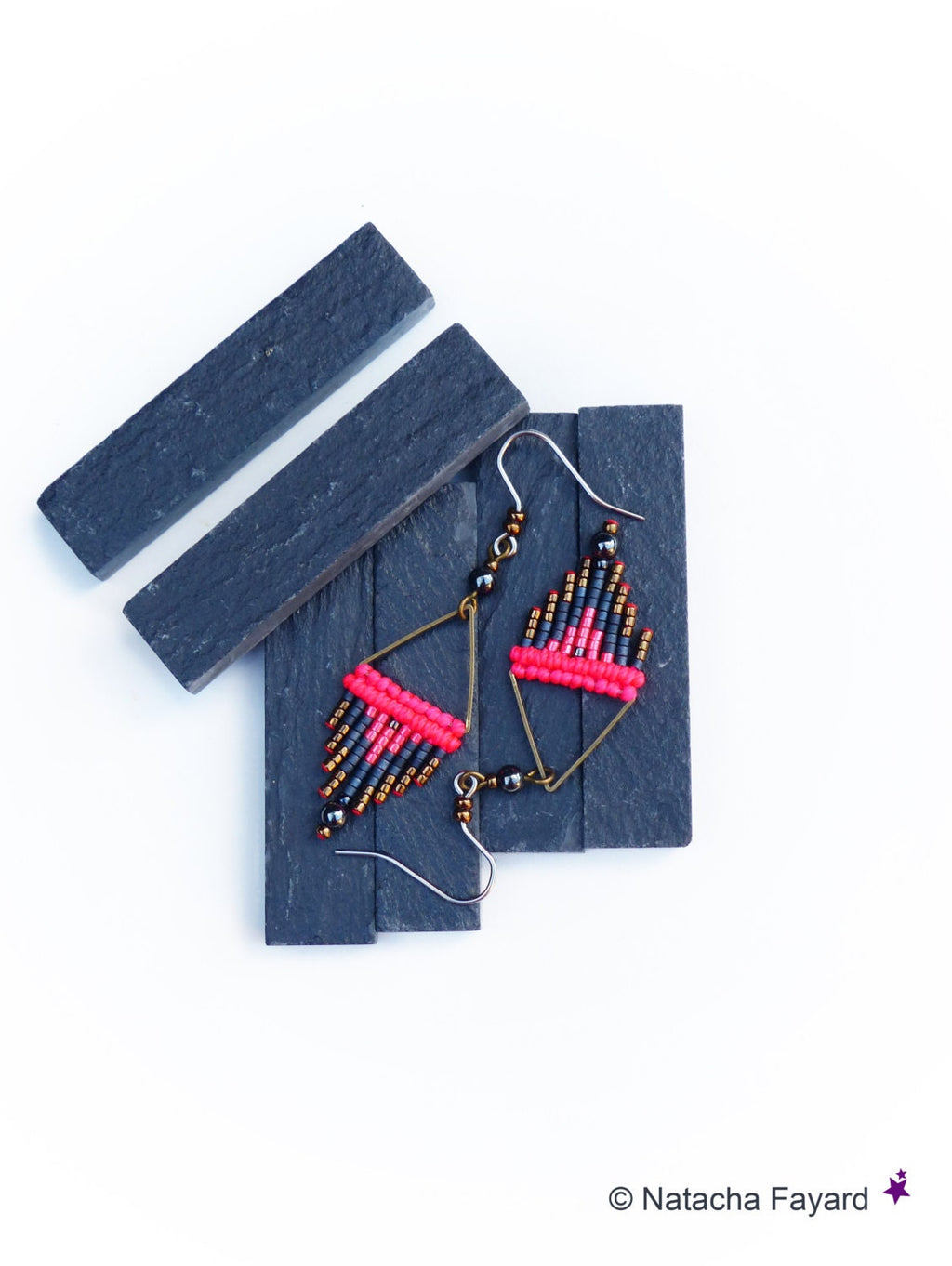 Peut inclure: Une paire de boucles d'oreilles pendantes avec un motif géométrique. Les boucles d'oreilles sont faites de perles noires, dorées et roses. Les boucles d'oreilles sont suspendues à une surface en ardoise noire.
