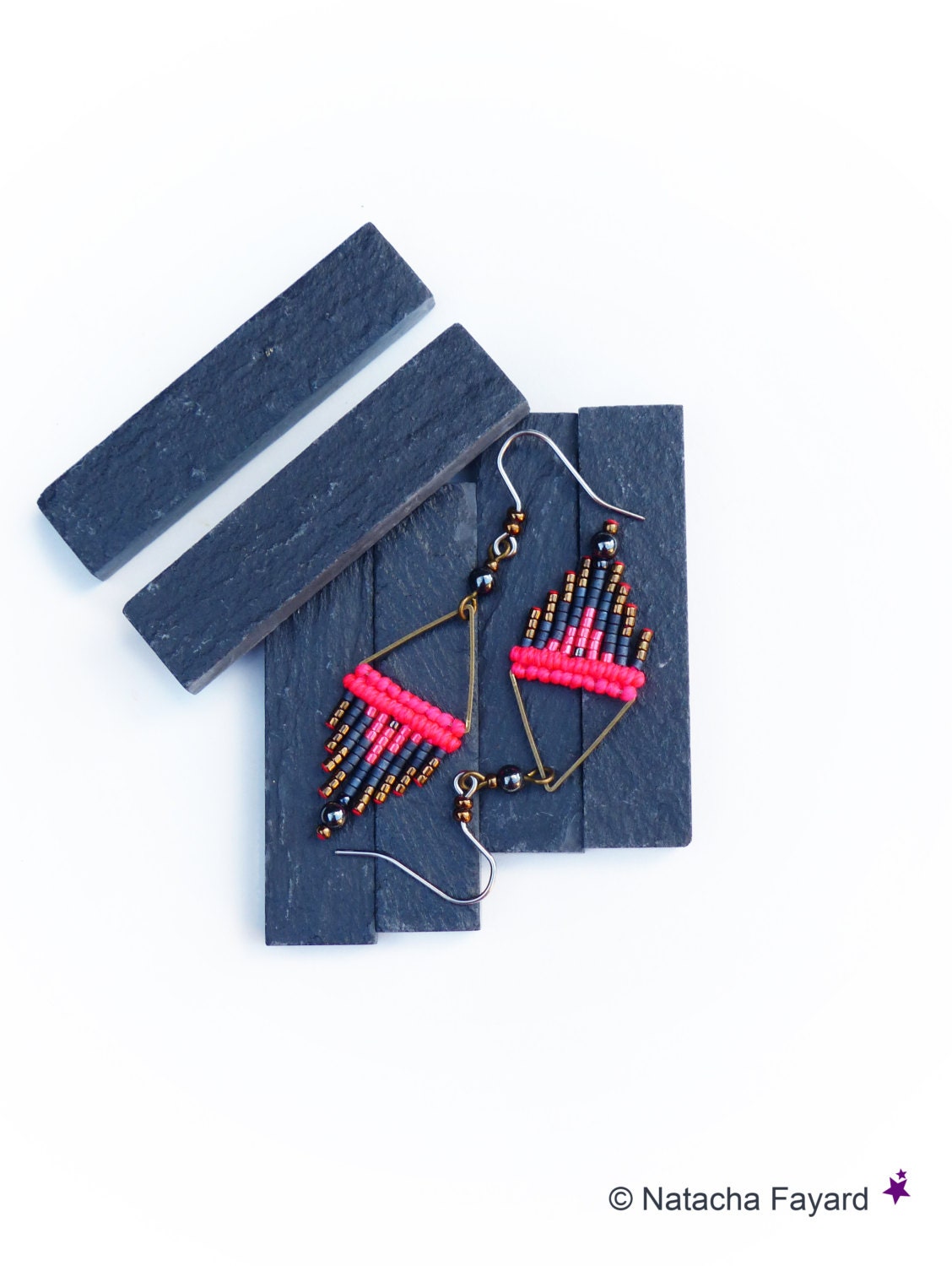 Peut inclure: Une paire de boucles d'oreilles pendantes avec un motif géométrique. Les boucles d'oreilles sont faites de perles noires, dorées et roses. Les boucles d'oreilles sont suspendues à une surface en ardoise noire.