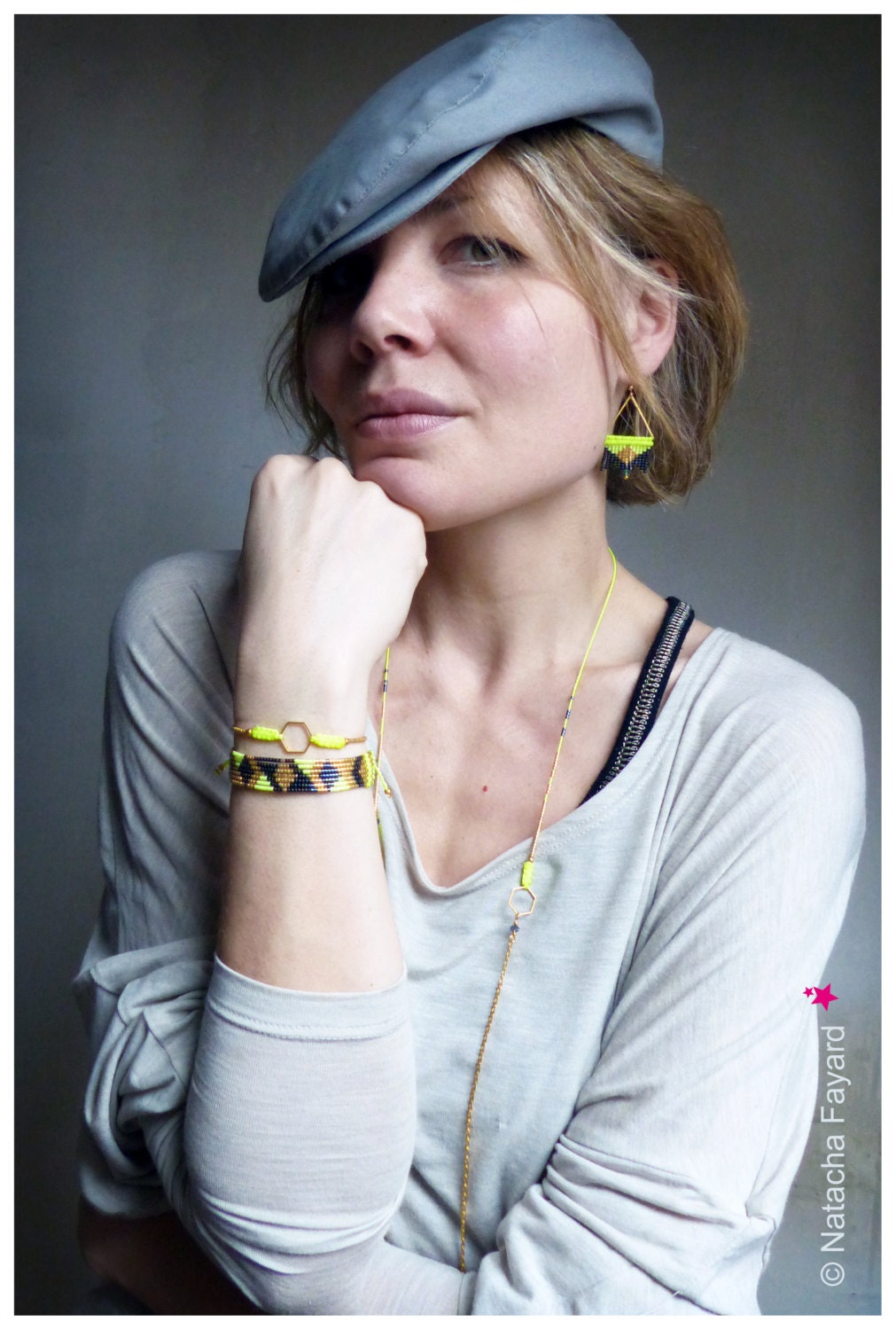 Peut inclure: Une femme portant une casquette plate grise, un t-shirt blanc et un collier en chaîne dorée avec une perle jaune. Elle porte également un bracelet en perles noires et jaunes et une paire de boucles d'oreilles pendantes jaunes et noires.