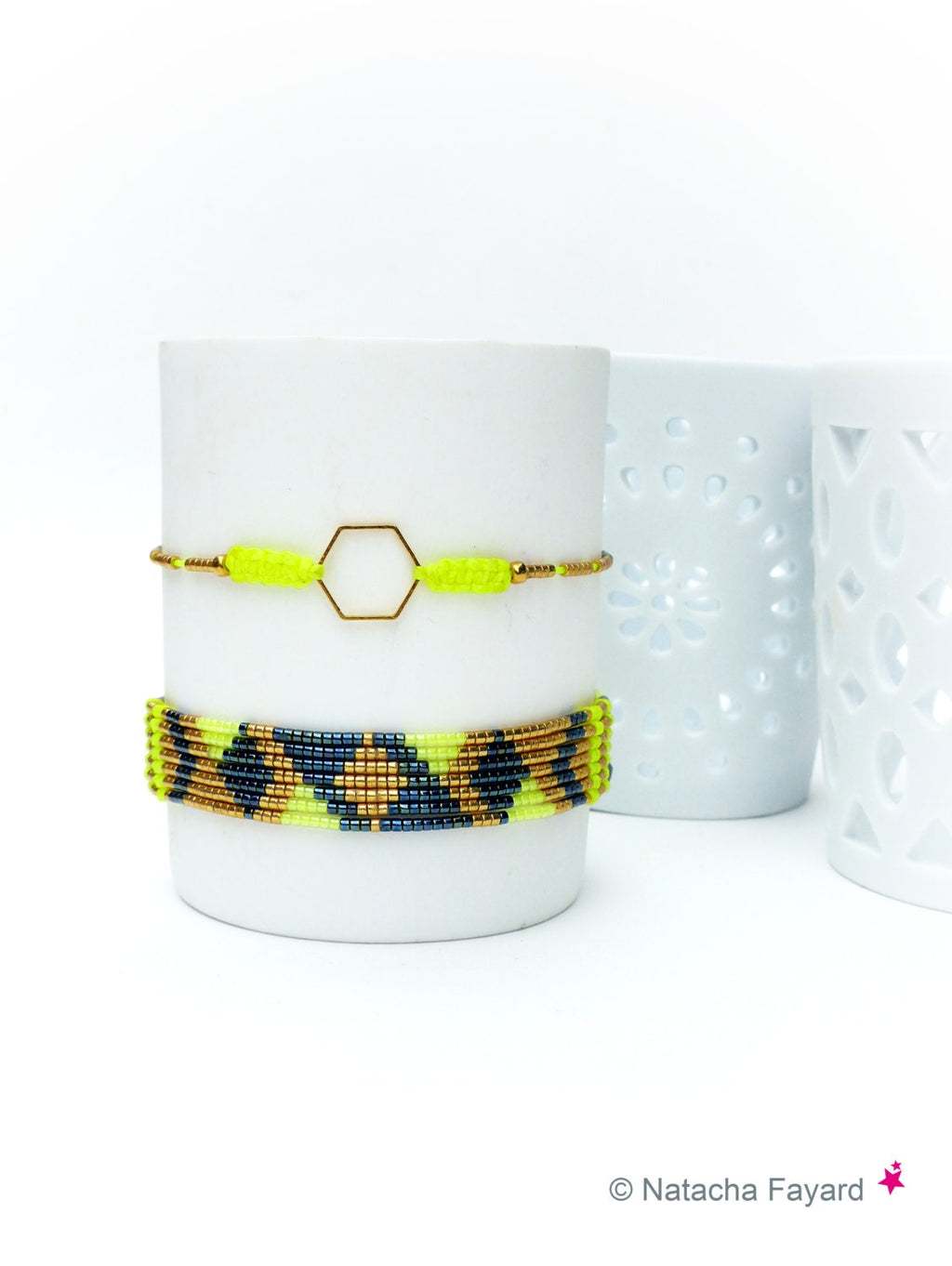 Peut inclure: Deux bracelets de perles. L'un présente une bande jaune fluo avec un hexagone doré et des perles dorées. L'autre bracelet a un motif de perles jaune fluo, bleu marine et dorées. Les deux bracelets sont exposés sur un objet cylindrique blanc.