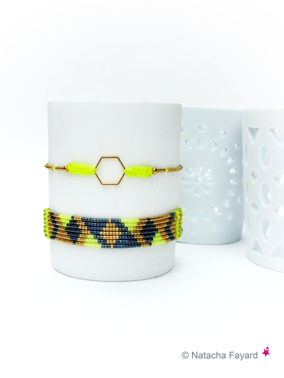 Peut inclure: Deux bracelets de perles. L'un présente une bande jaune fluo avec un hexagone doré et des perles dorées. L'autre bracelet a un motif de perles jaune fluo, bleu marine et dorées. Les deux bracelets sont exposés sur un objet cylindrique blanc.