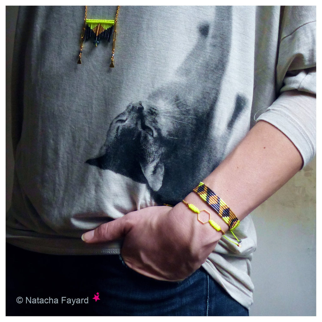 Peut inclure: Un t-shirt gris à manches longues avec un grand motif de chat noir et blanc. La personne porte deux bracelets : un avec un charm hexagonal jaune et l'autre avec un motif tissé noir et jaune. Un collier avec un design de perles jaunes et noires est également visible.