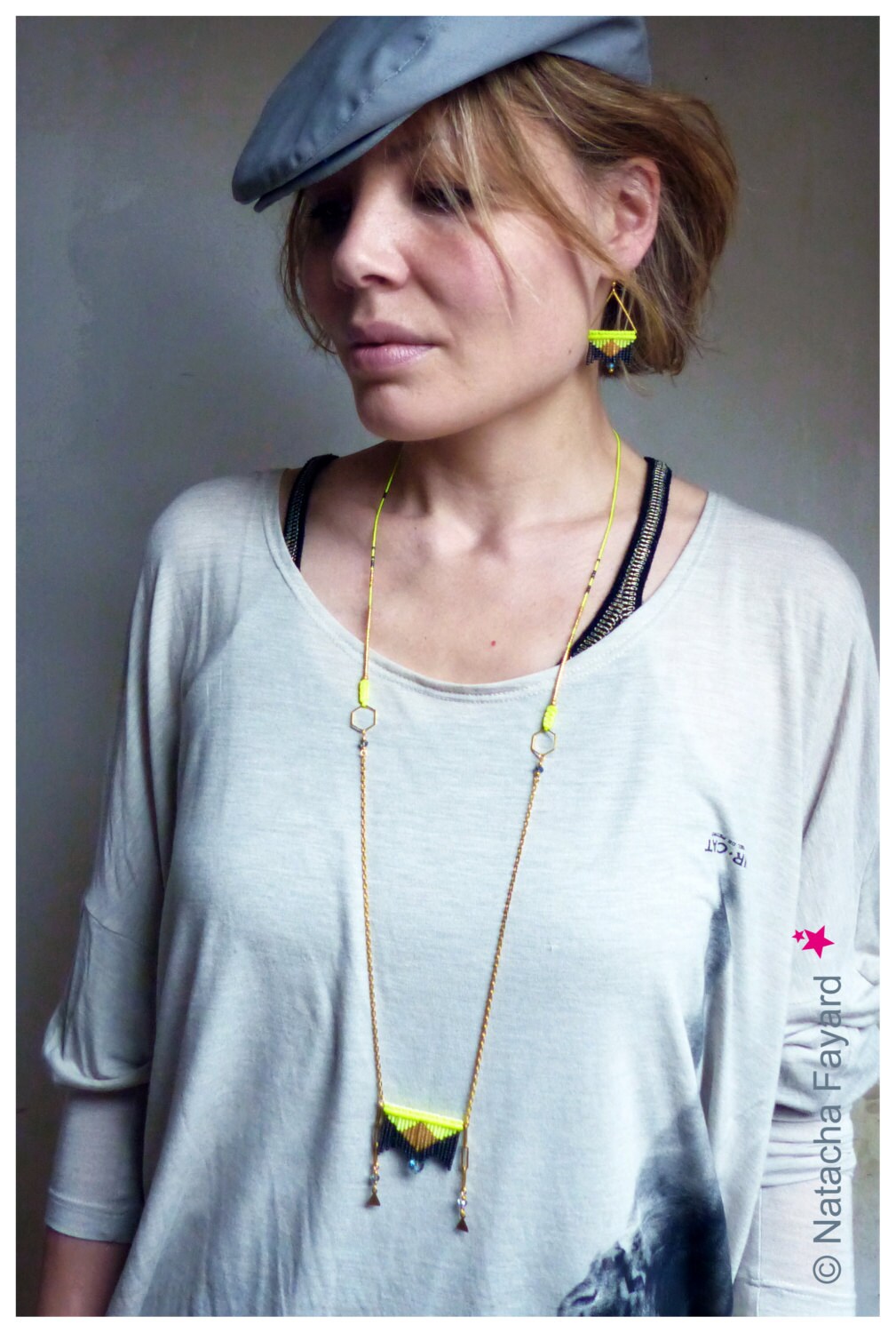 Peut inclure: Une femme portant une casquette grise et un t-shirt blanc avec un collier en chaîne dorée orné d'un pendentif géométrique aux accents noirs et jaunes. Le pendentif est de forme triangulaire avec une frange de perles noires et jaunes.