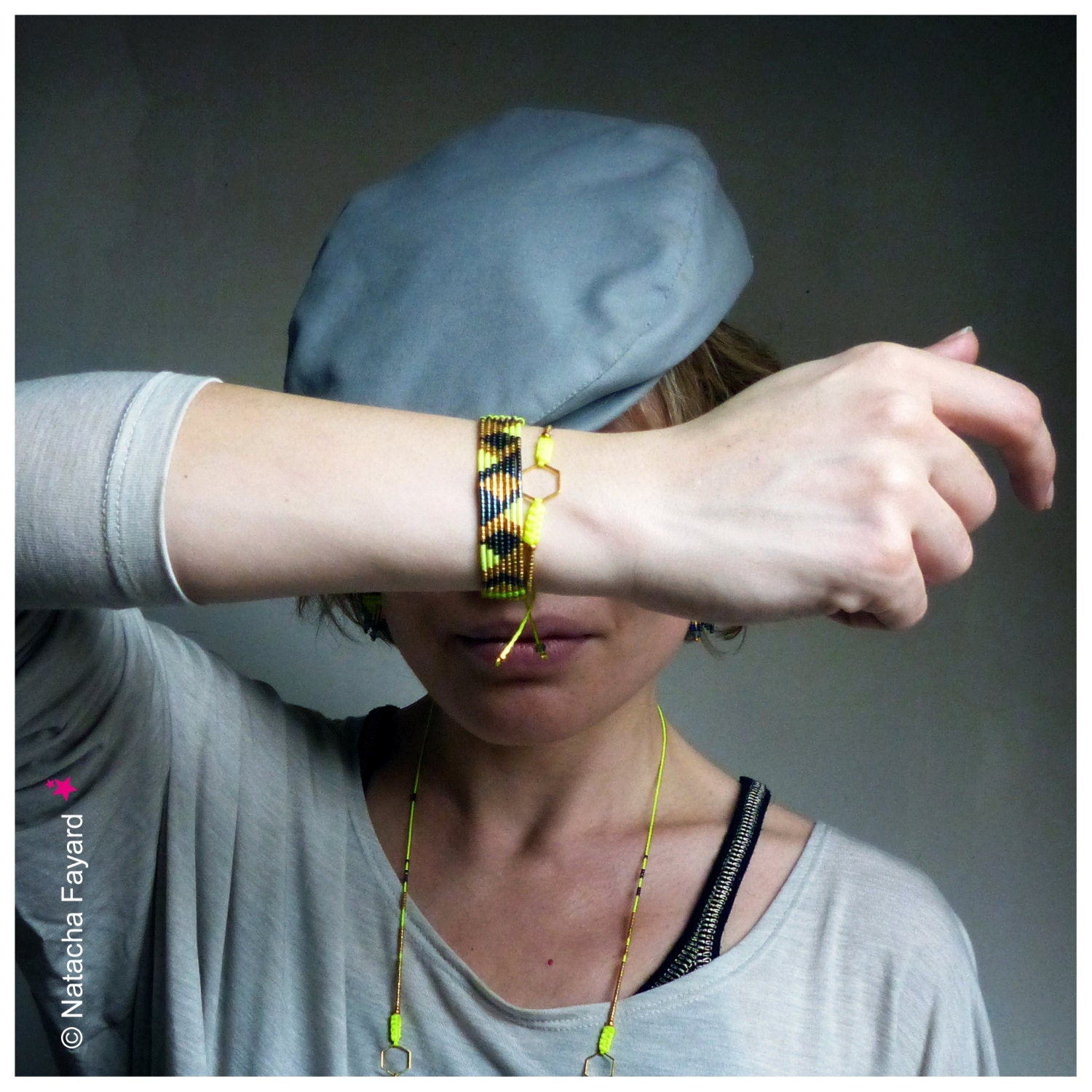 Peut inclure: Un bracelet de perles avec un motif géométrique noir, jaune et vert. Le bracelet est porté au poignet, avec un cordon jaune assorti et un charm doré. Un béret gris est visible en arrière-plan.