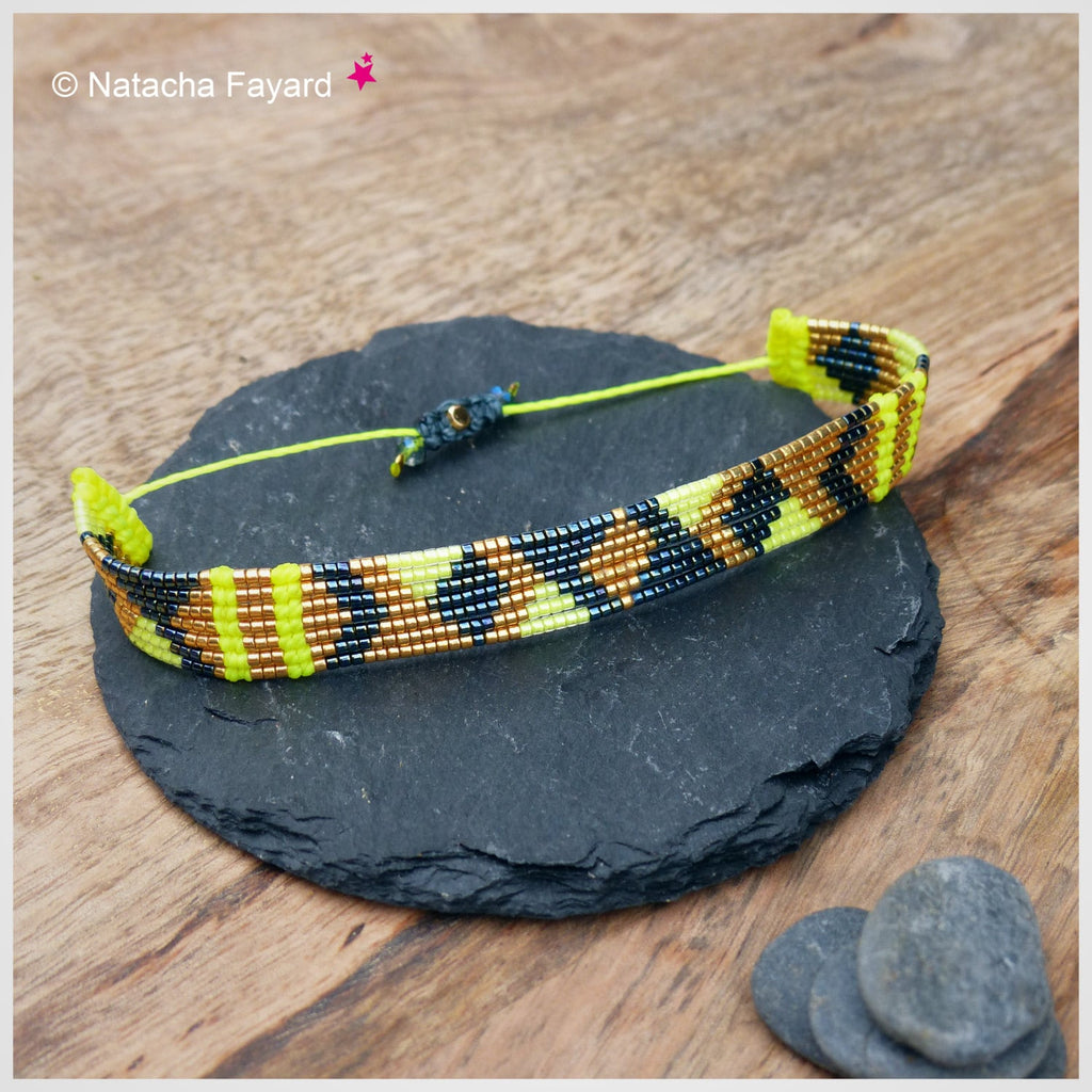 Peut inclure: Un bracelet de perles avec un motif géométrique jaune, noir et marron. Le bracelet est posé sur une surface en pierre grise foncée. Il est composé de petites perles et possède une fermeture à cordon jaune. Le texte "Natacha Fayard" est visible.