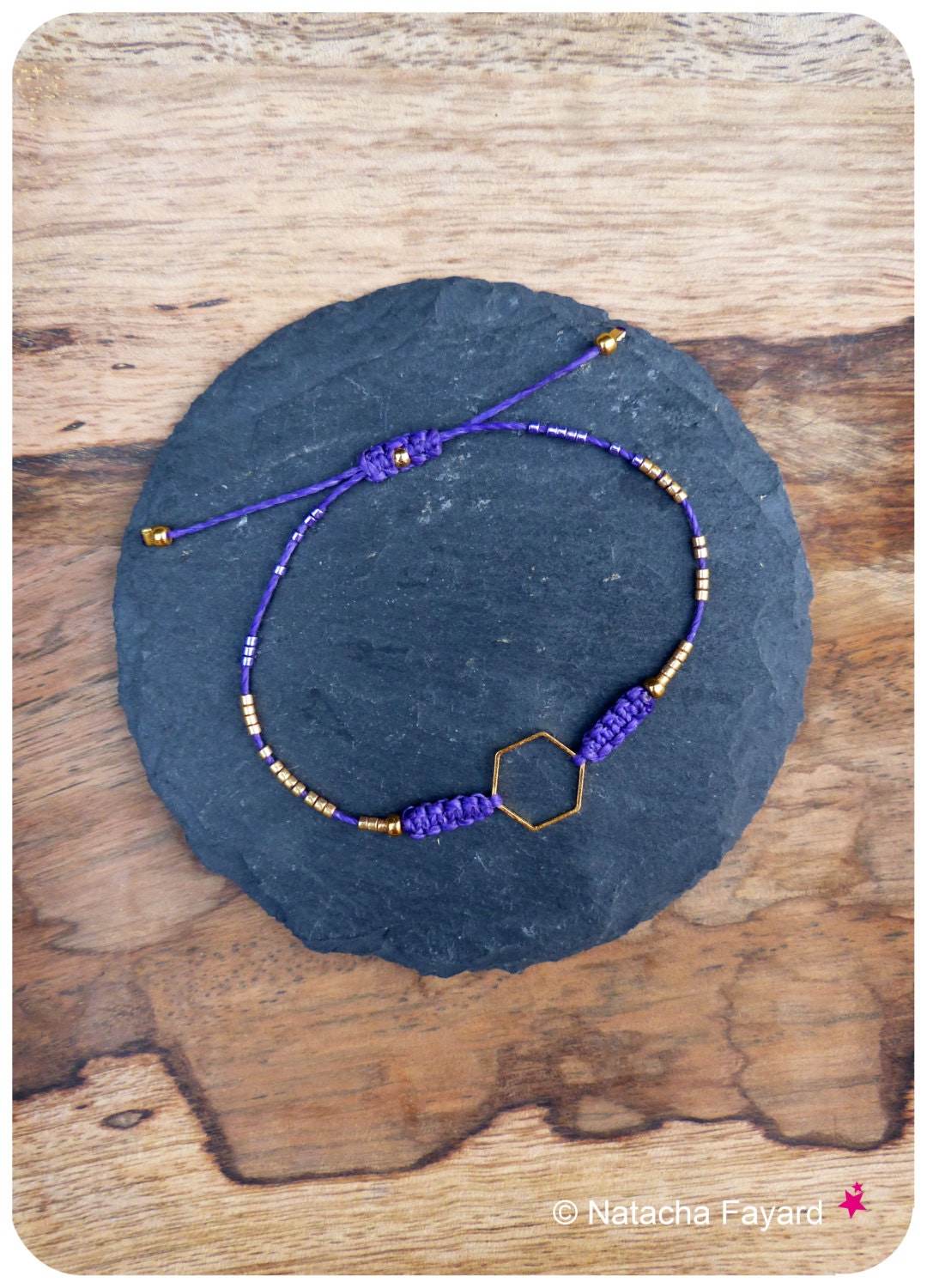 Peut inclure: Un bracelet en cordon violet doté d'une breloque hexagonale dorée centrale, encadrée par deux nœuds en macramé violets et agrémentée de petites perles métalliques dorées réparties le long du cordon, avec un fermoir coulissant réglable.