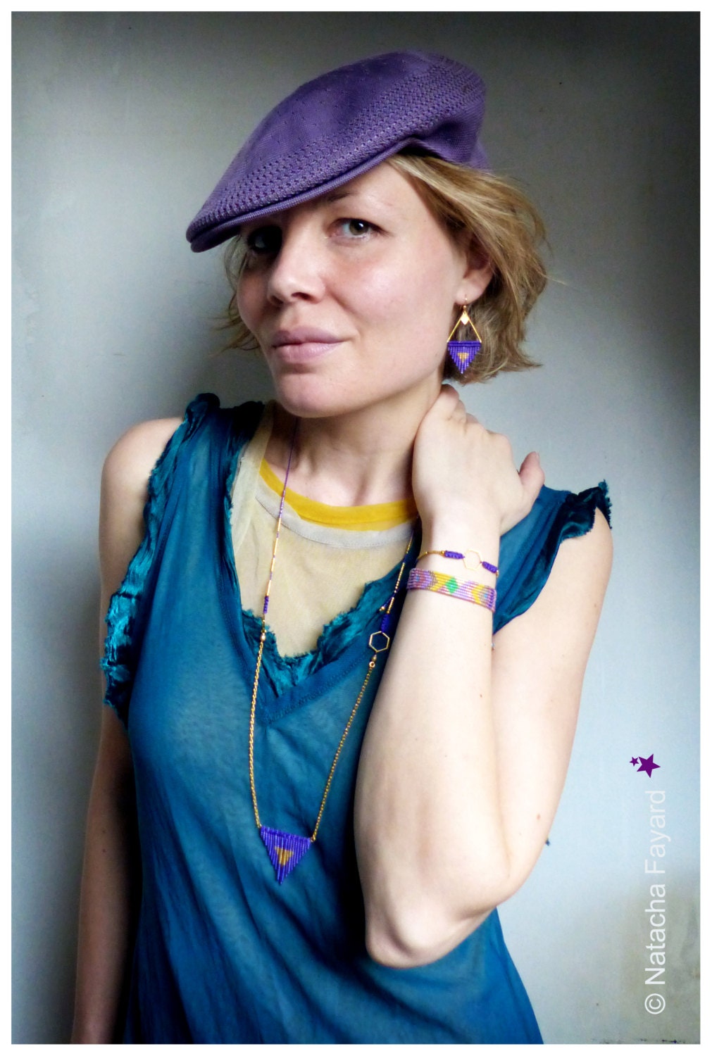 Peut inclure: Un bonnet en tricot violet, un long collier à chaîne dorée avec un pendentif triangle violet, une boucle d'oreille triangle violette assortie et un bracelet tissé à motif géométrique violet, jaune et blanc. Le texte Natacha Fayard est visible.