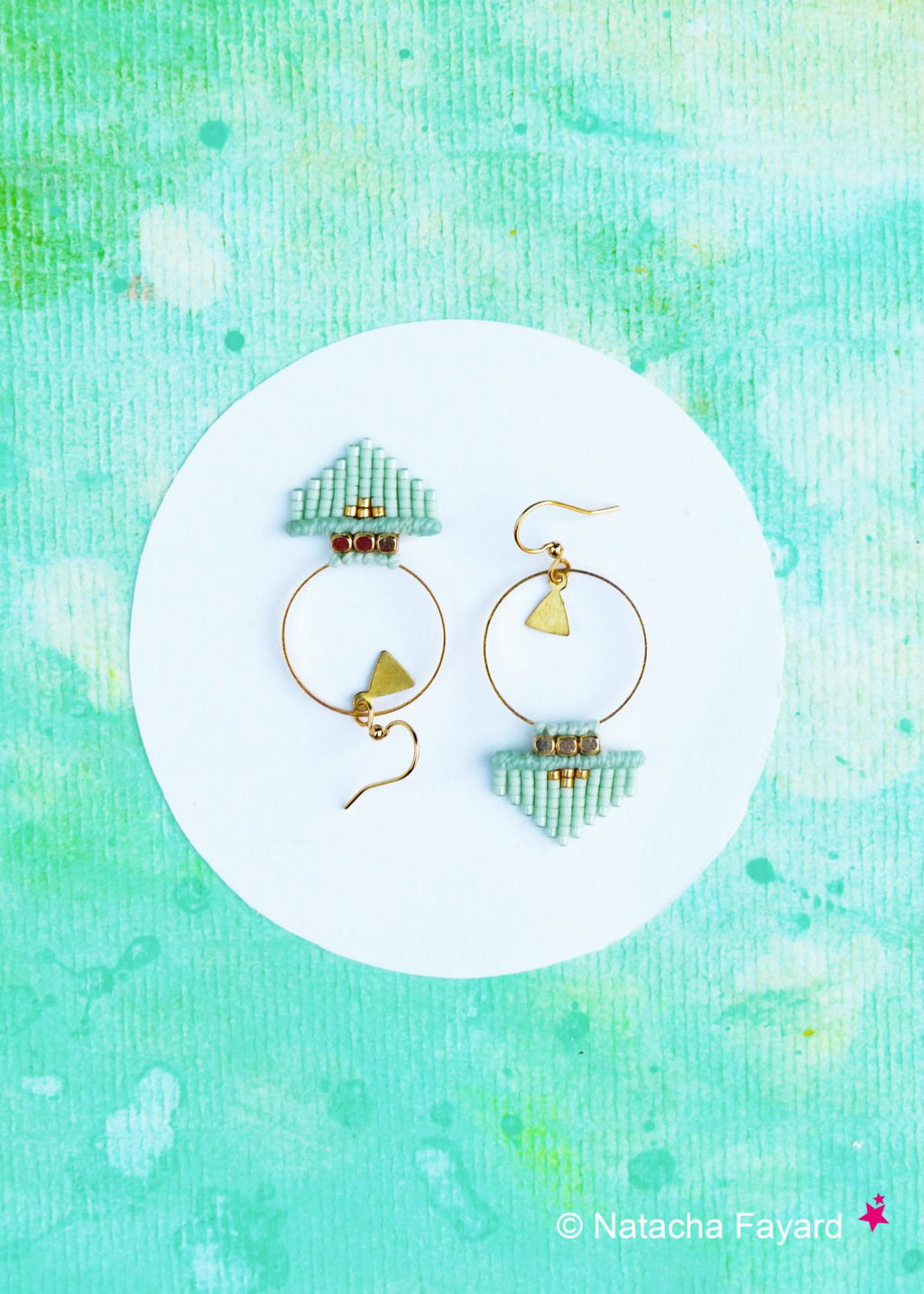 Peut inclure: Une paire de boucles d'oreilles en perles vert clair avec des cercles dorés et des breloques triangulaires. Les boucles d'oreilles sont sur un fond blanc.